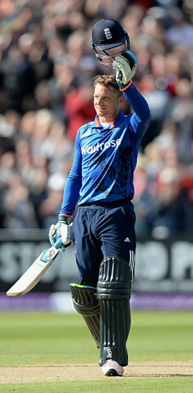 jos buttler 桌布 壁紙 — 一名身穿藍色球衣的板球運動員在陽光明媚的球場上慶祝，高舉球棒和頭盔。