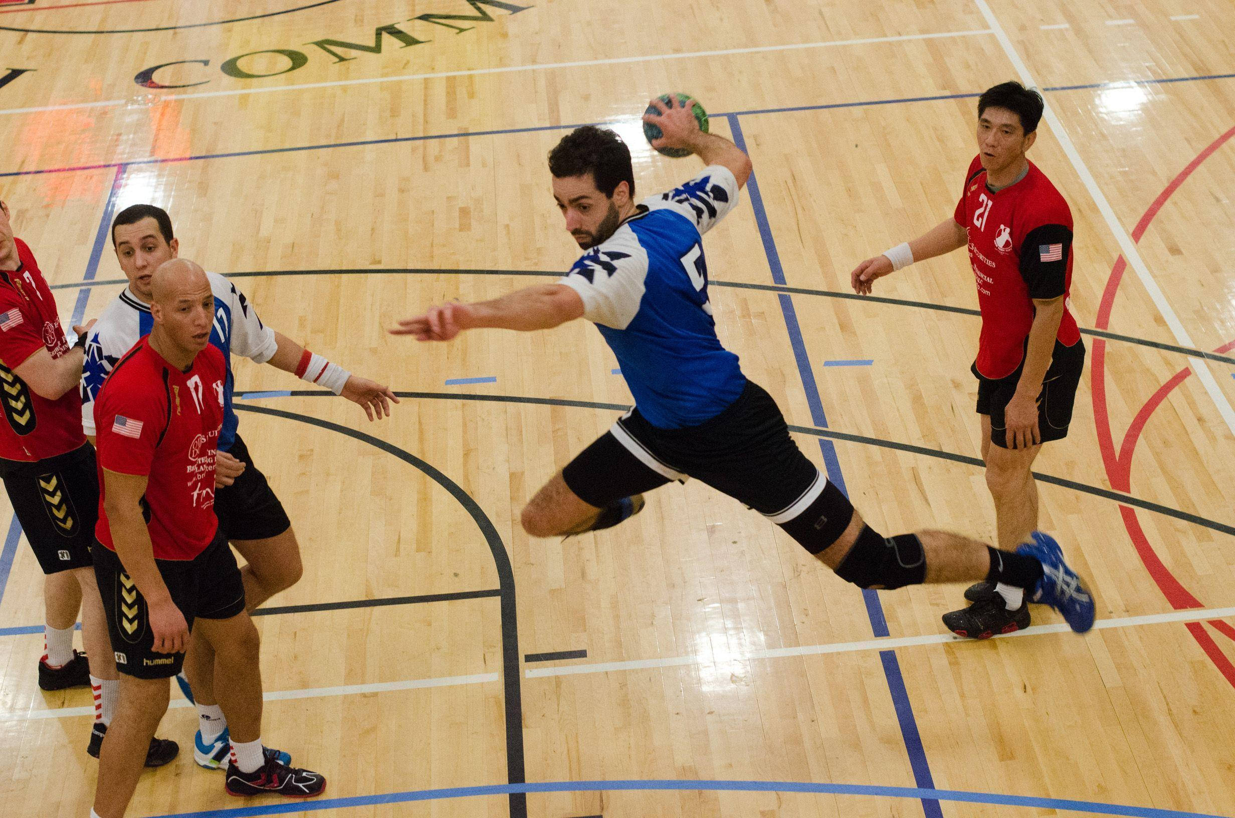 手球 桌布 壁紙 — 一名身穿藍色球衣的男子手球運動員在拋光木地板上動態跳躍投籃，而紅色防守球員正逼近。