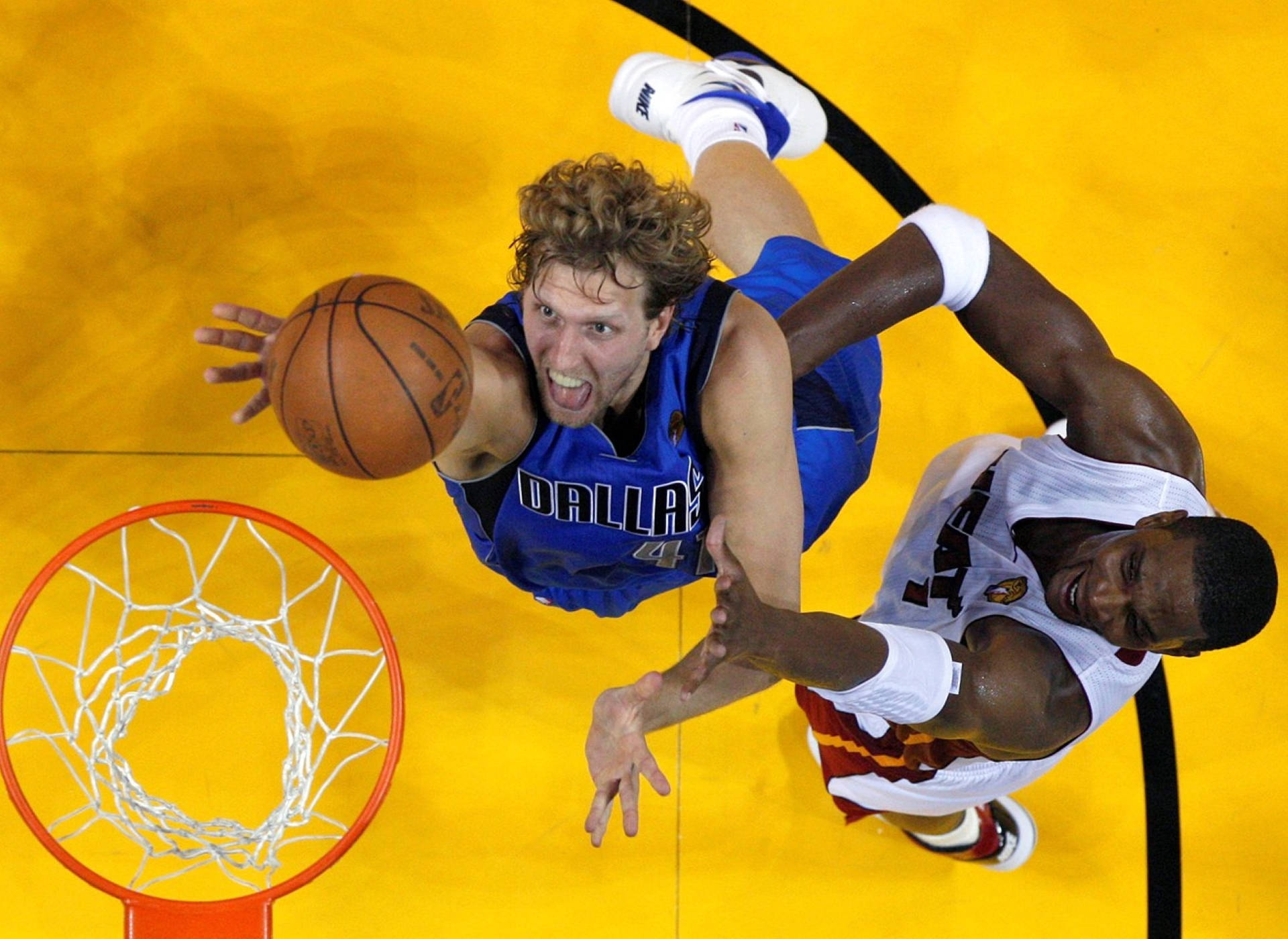 Dirk Nowitzki 桌布 壁紙 — 一名身穿藍色球衣的籃球運動員向籃框飛撲，而一名身穿白色球衣的防守球員試圖封阻他的投籃。