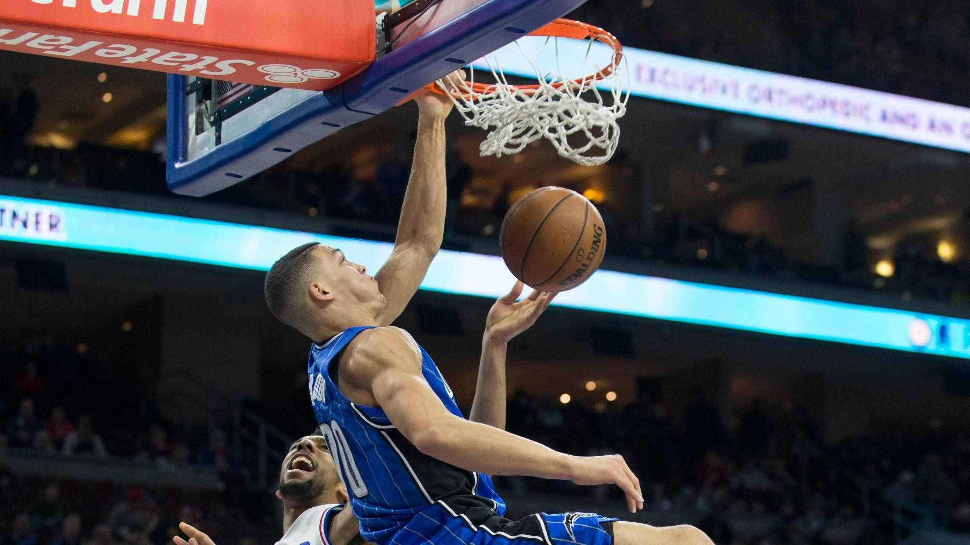 aaron gordon 桌布 壁紙 — 一名身穿藍色球衣的籃球運動員在燈光明亮的場館中，在防守球員下方觀看下，專注地灌籃。