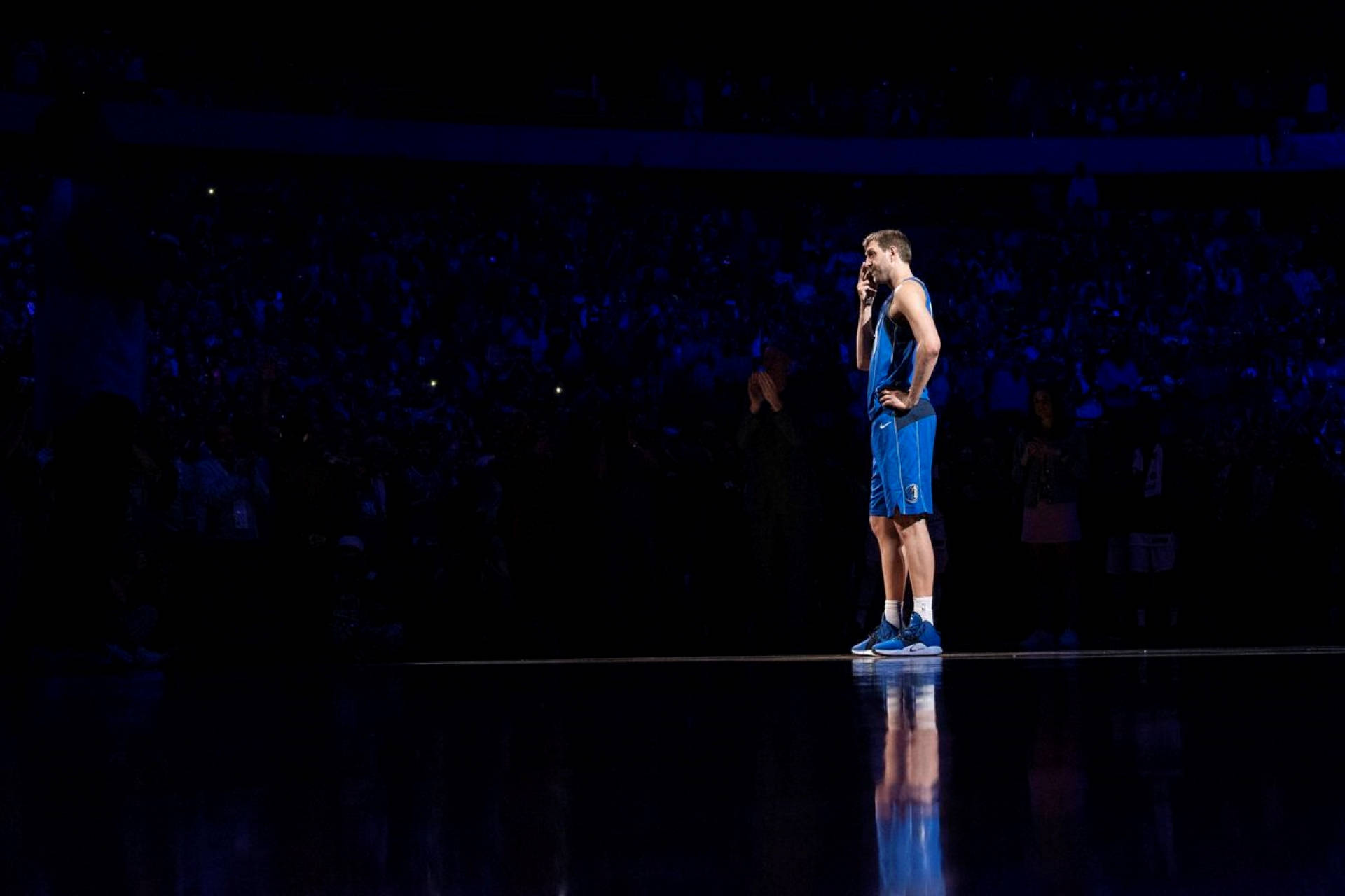 Dirk Nowitzki 桌布 壁紙 — 一名身穿藍色球衣的籃球運動員獨自站在聚光燈下的黑暗球場上，背景有觀眾。