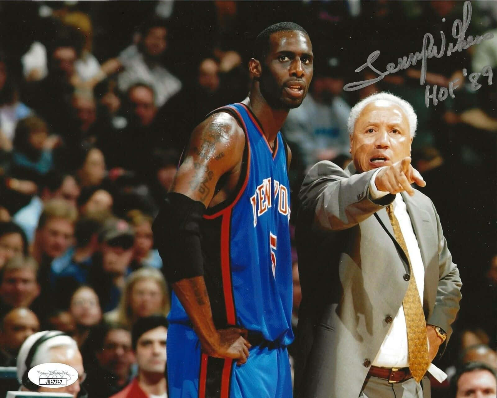 Coach 桌布 壁紙 — 一名身穿藍色球衣的籃球運動員站在一位身穿西裝的教練旁邊，教練在比賽中專注地指點，背後是模糊的人群。