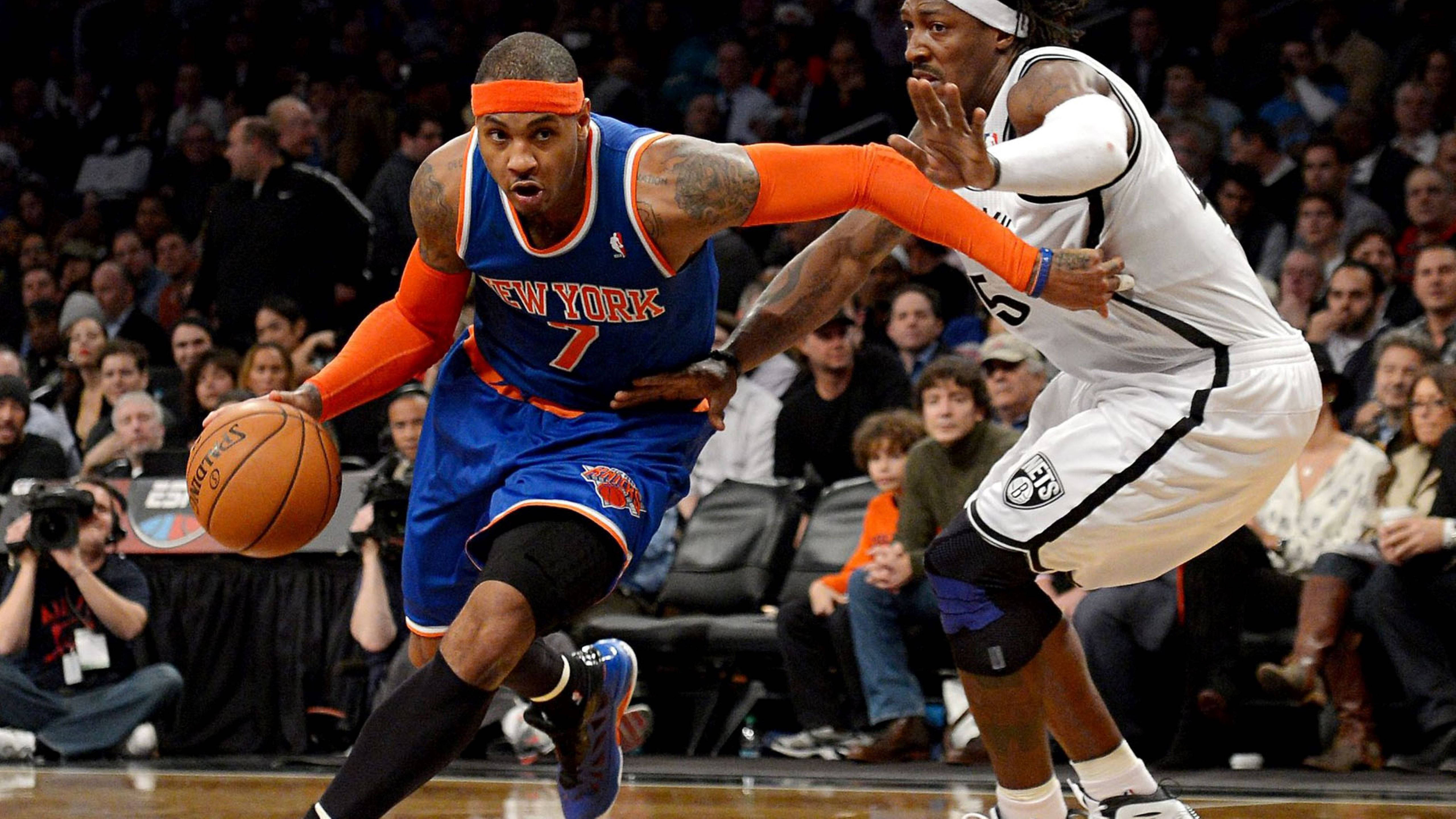 Carmelo 桌布 壁紙 — 一名身穿藍色球衣的紐約尼克球員在擠滿觀眾的場館中，運球越過布魯克林籃網隊的防守球員，進行一場高能量的籃球比賽。