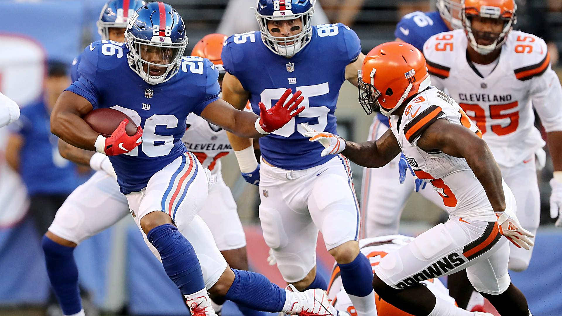 Saquon Barkley 桌布 壁紙 — 一名身穿藍色球衣的紐約巨人隊跑衛，在陽光明媚的球場上持球衝刺，身後有克里夫蘭布朗隊的防守球員追趕。