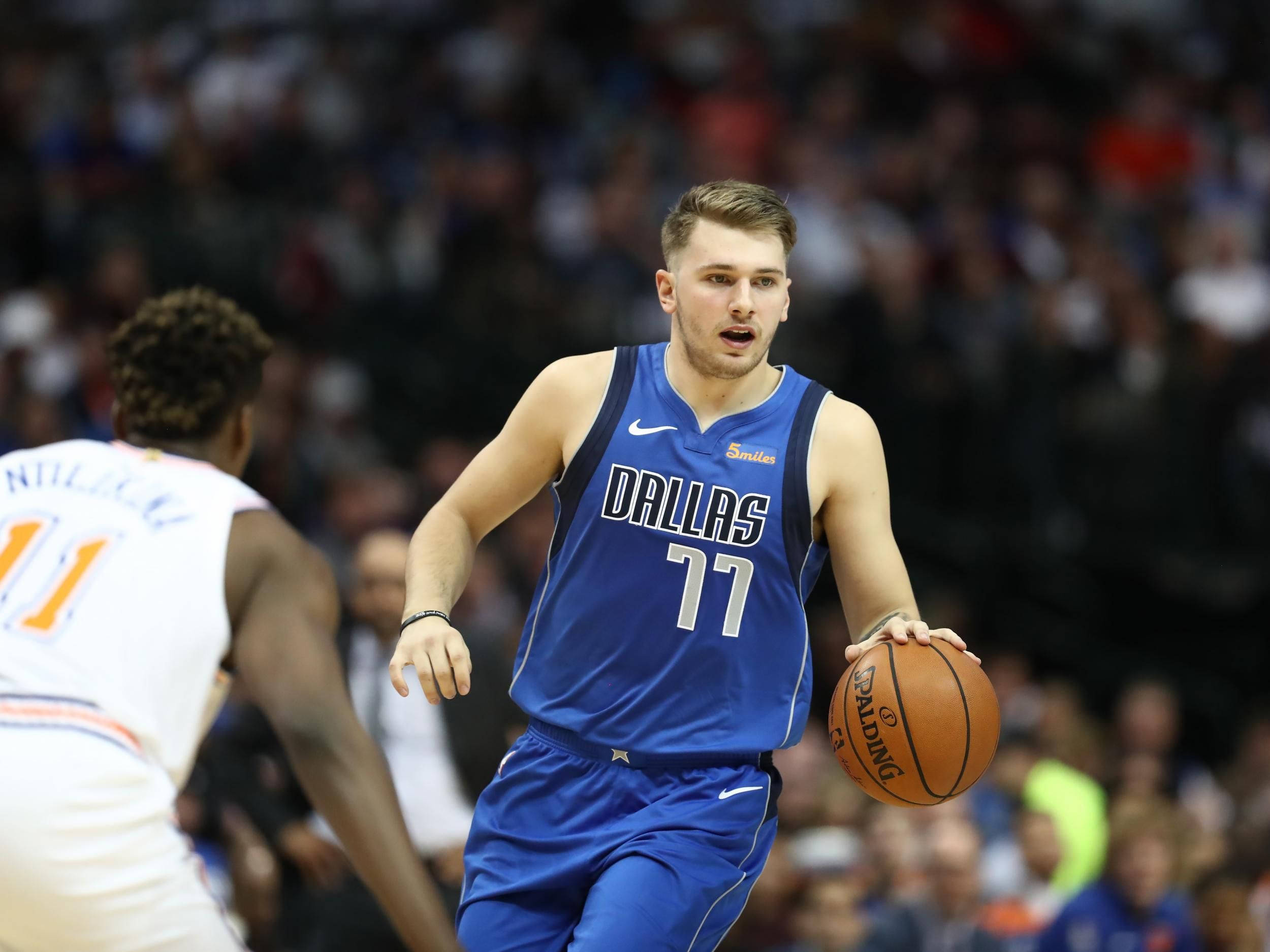 Luka 桌布 壁紙 — 一名身穿藍色球衣的達拉斯獨行俠籃球球員在激烈的比賽中運球，背景是模糊的人群。