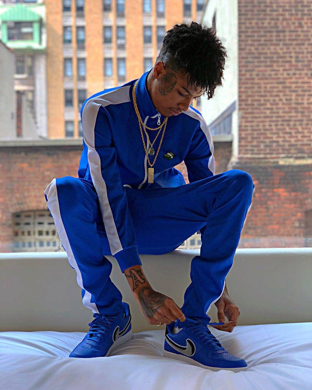 Celebrity 桌布 壁紙 — 一名身穿藍色白條紋運動服的年輕男子坐在床上，背後是城市天際線，正在繫他相配的藍色運動鞋鞋帶。