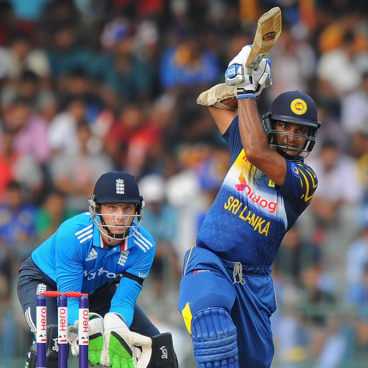 Sri Lanka Cricket 桌布 壁紙 — 一名身穿藍色的斯里蘭卡板球擊球手強力揮棒，而一名英格蘭守門員警覺地蹲在門柱後面。