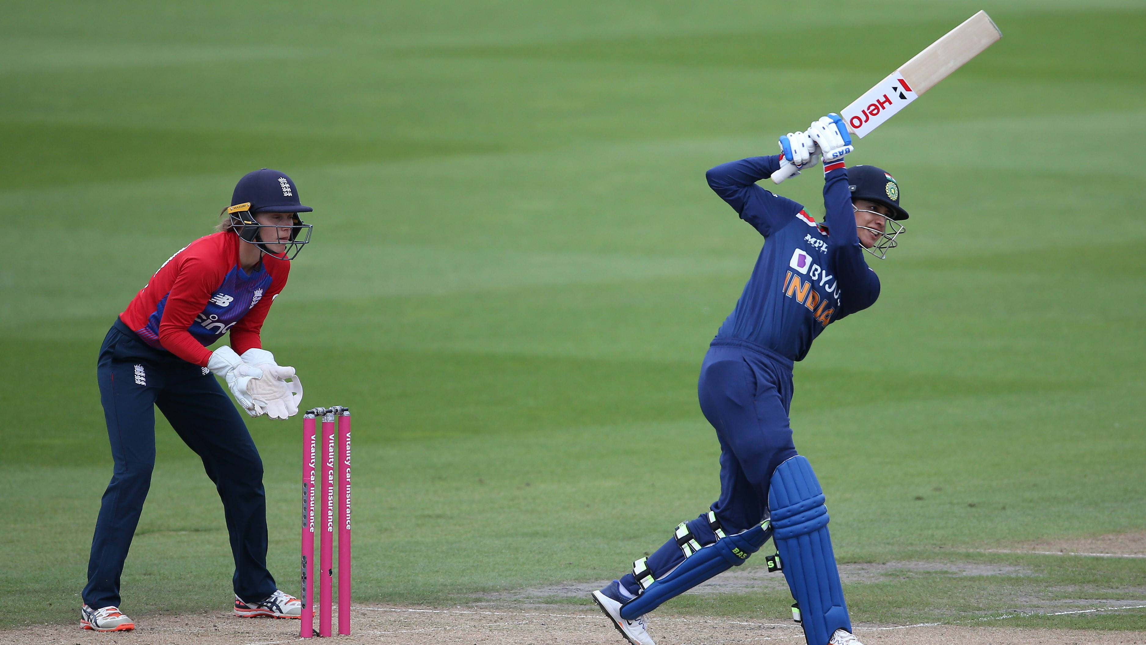 Smriti Mandhana 桌布 壁紙 — 一名身穿藍色的板球擊球手強力揮擊球，而一名身穿紅色的守門員蹲在粉紅色三柱門後，在充滿活力的綠色場地上。