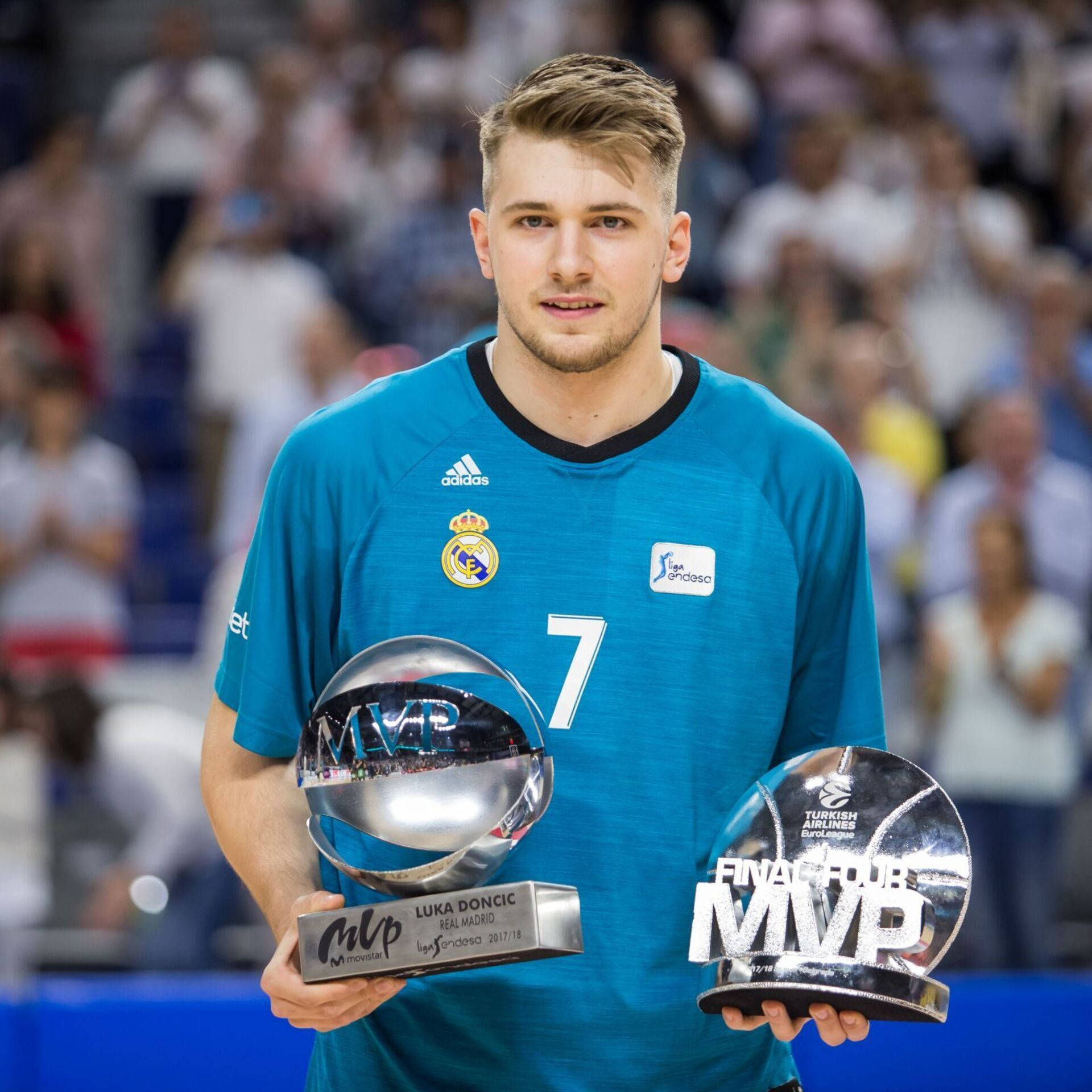 Luka 桌布 壁紙 — 一名身穿藍色皇家馬德里球衣的籃球運動員，在模糊的人群背景中站在球場上，手持兩個閃亮的MVP獎盃。