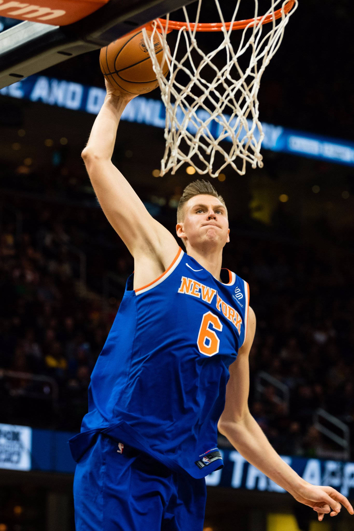 Kristaps Porzingis 桌布 壁紙 — 一名身穿藍色紐約尼克球衣的籃球運動員在明亮的場館燈光下，以高度專注力灌籃。