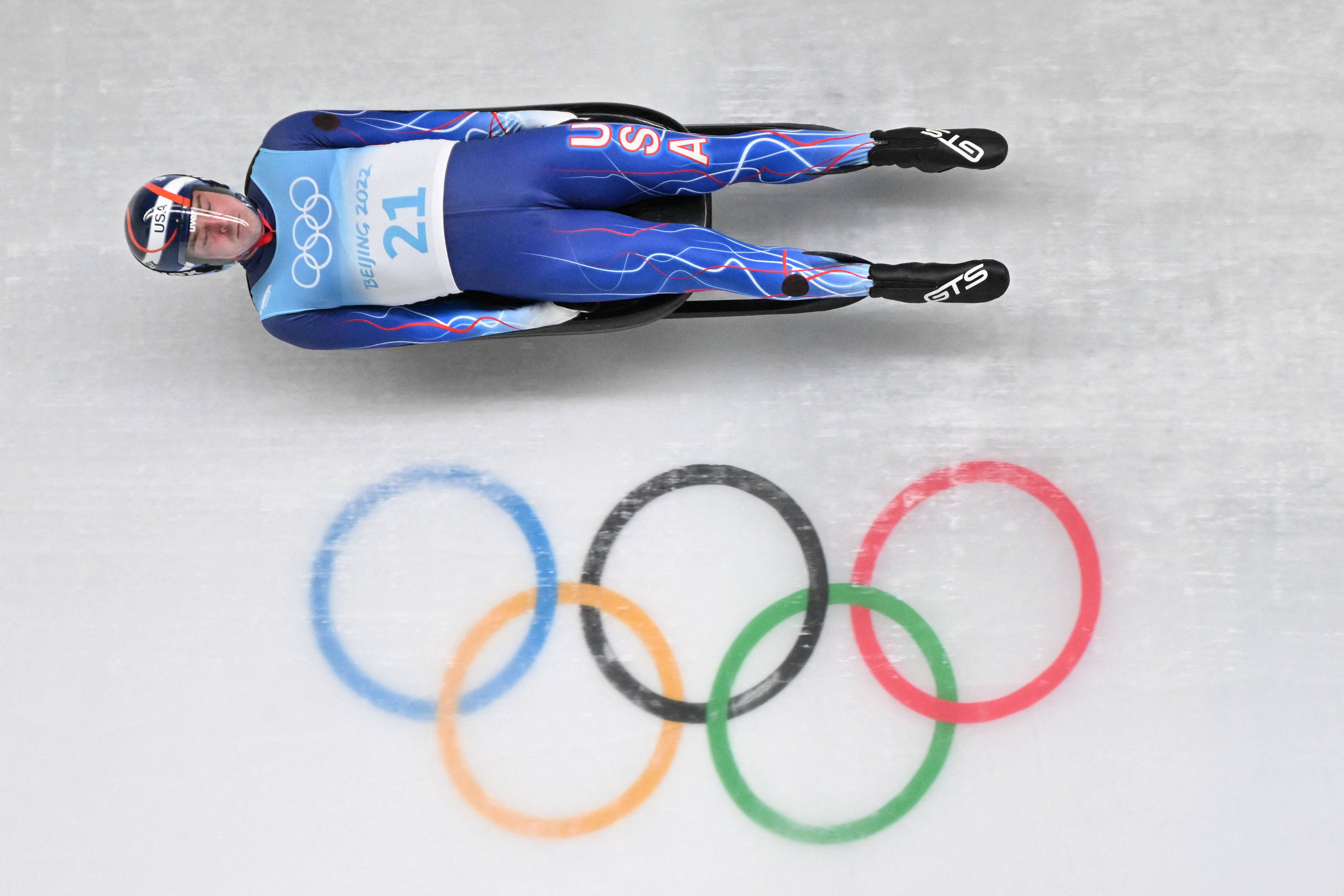 Athletic 桌布 壁紙 — 一名身穿藍色美國隊服的雪橇運動員在冰道上飛馳，下方可見奧運五環。