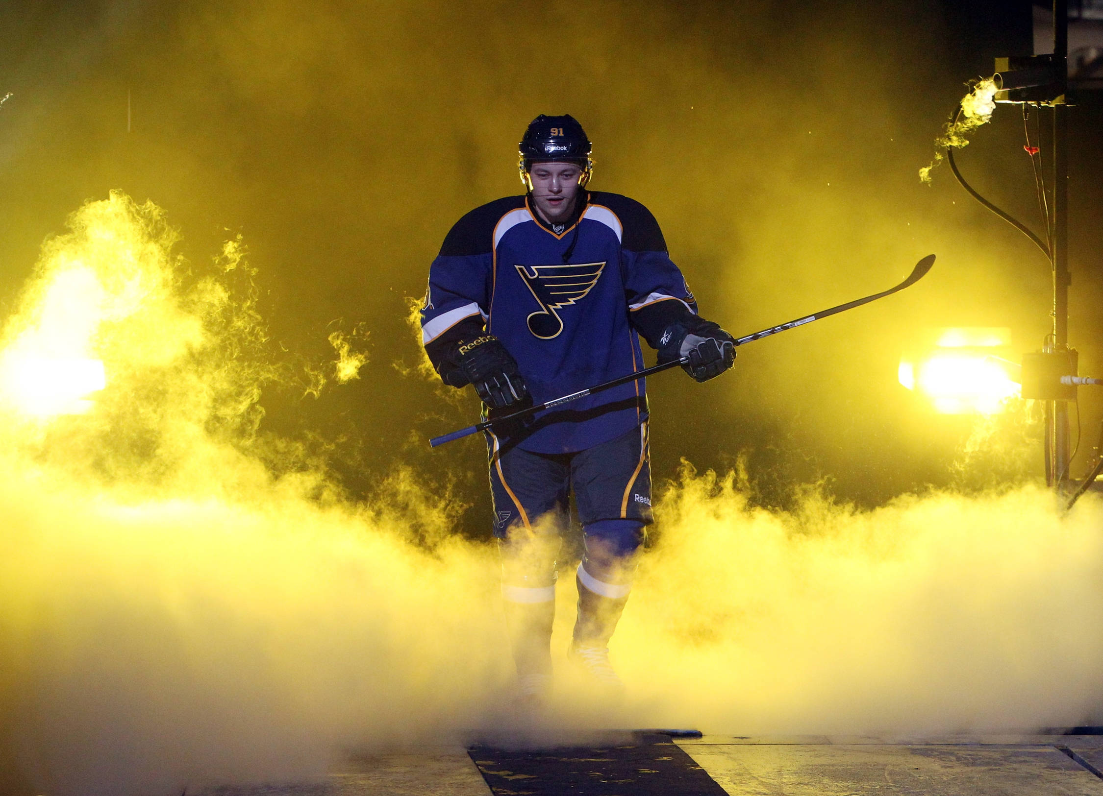 St Louis Blues 桌布 壁紙 — 一名身穿藍色聖路易斯藍調球衣的冰球運動員從黃色煙霧中出現，在戲劇性的場館燈光下拿著球桿。