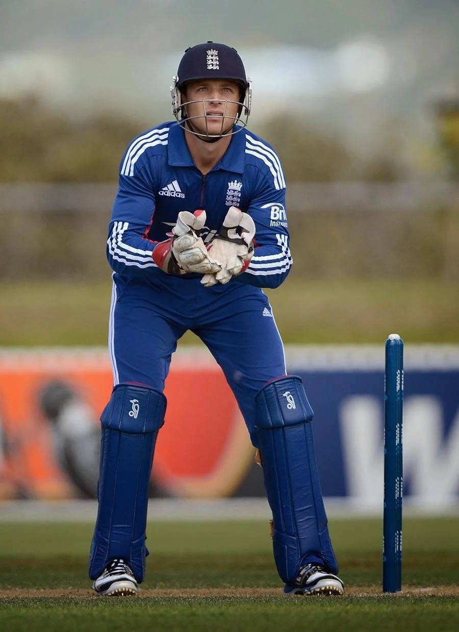 jos buttler 桌布 壁紙 — 一名身穿藍色英格蘭球衣的板球擊球手在草地上警覺地蹲在球門附近。