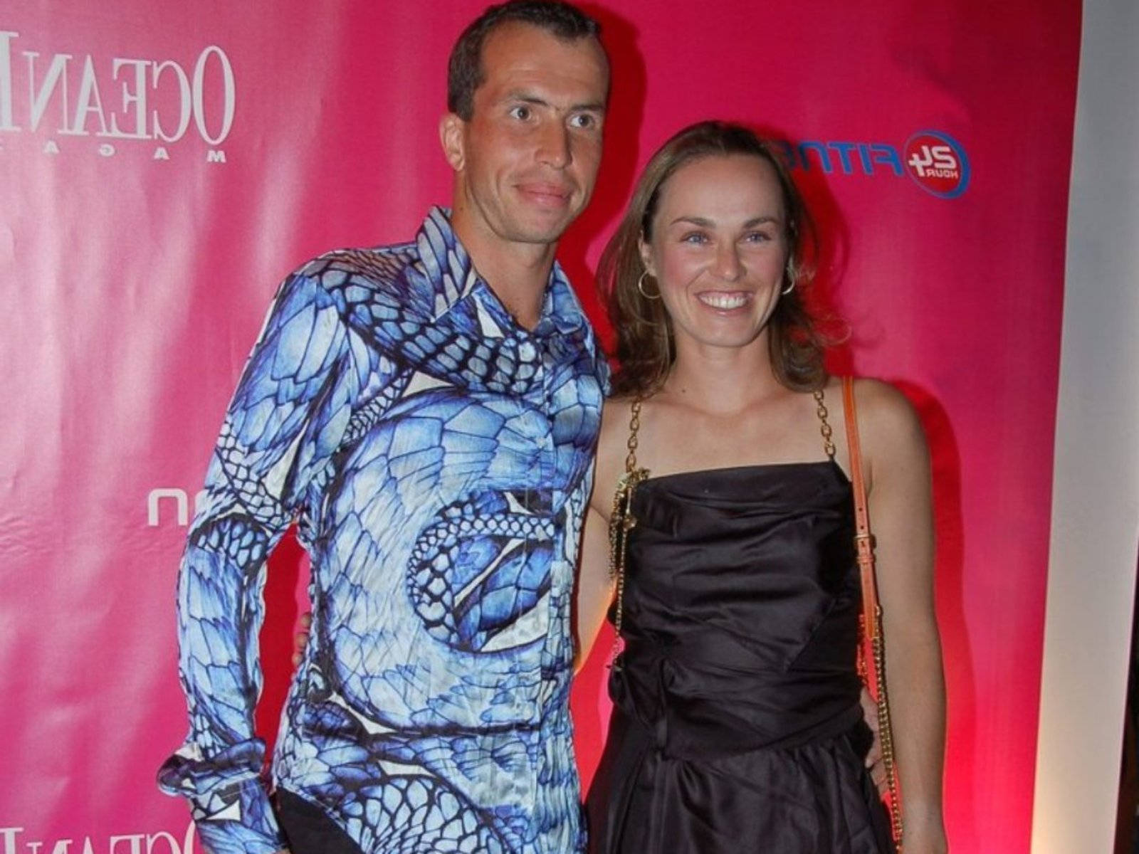 martina hingis 桌布 壁紙 — 一名身穿藍色蛇紋襯衫的男子和一名身穿黑色連衣裙的女子在粉紅色品牌背景前微笑拍照。