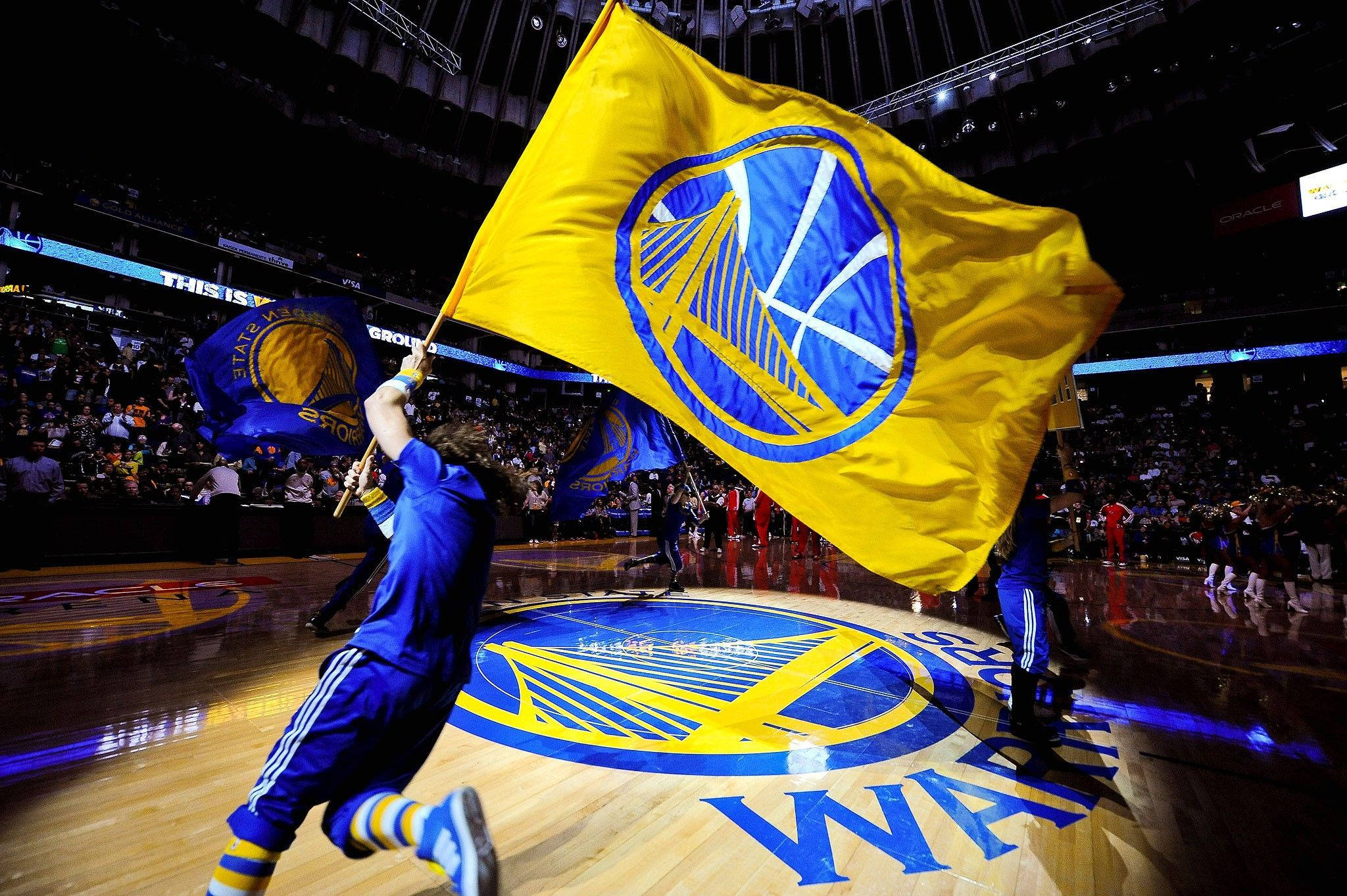 Warriors 桌布 壁紙 — 一名身穿藍色衣服的球迷在比賽期間跑過籃球場，揮舞著一面大的金色金州勇士隊旗幟。