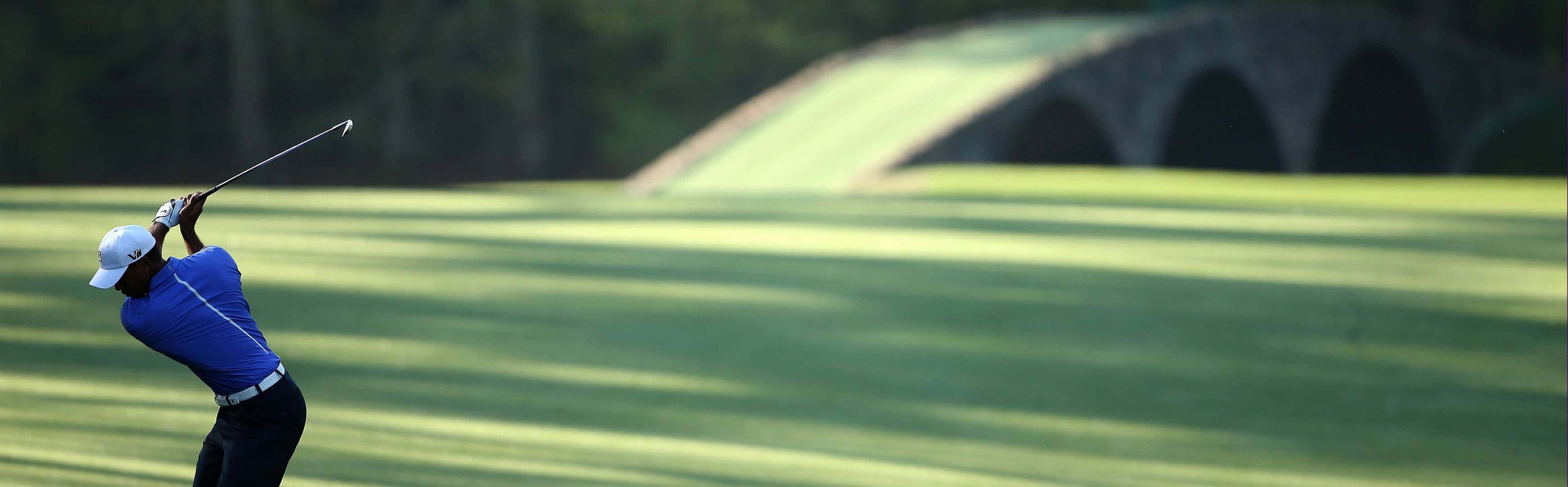 Golf 3840x1080 桌布 壁紙 — 一名身穿藍色衣服的高爾夫球手在陽光明媚的球道上強力揮桿，背景是一座石橋。