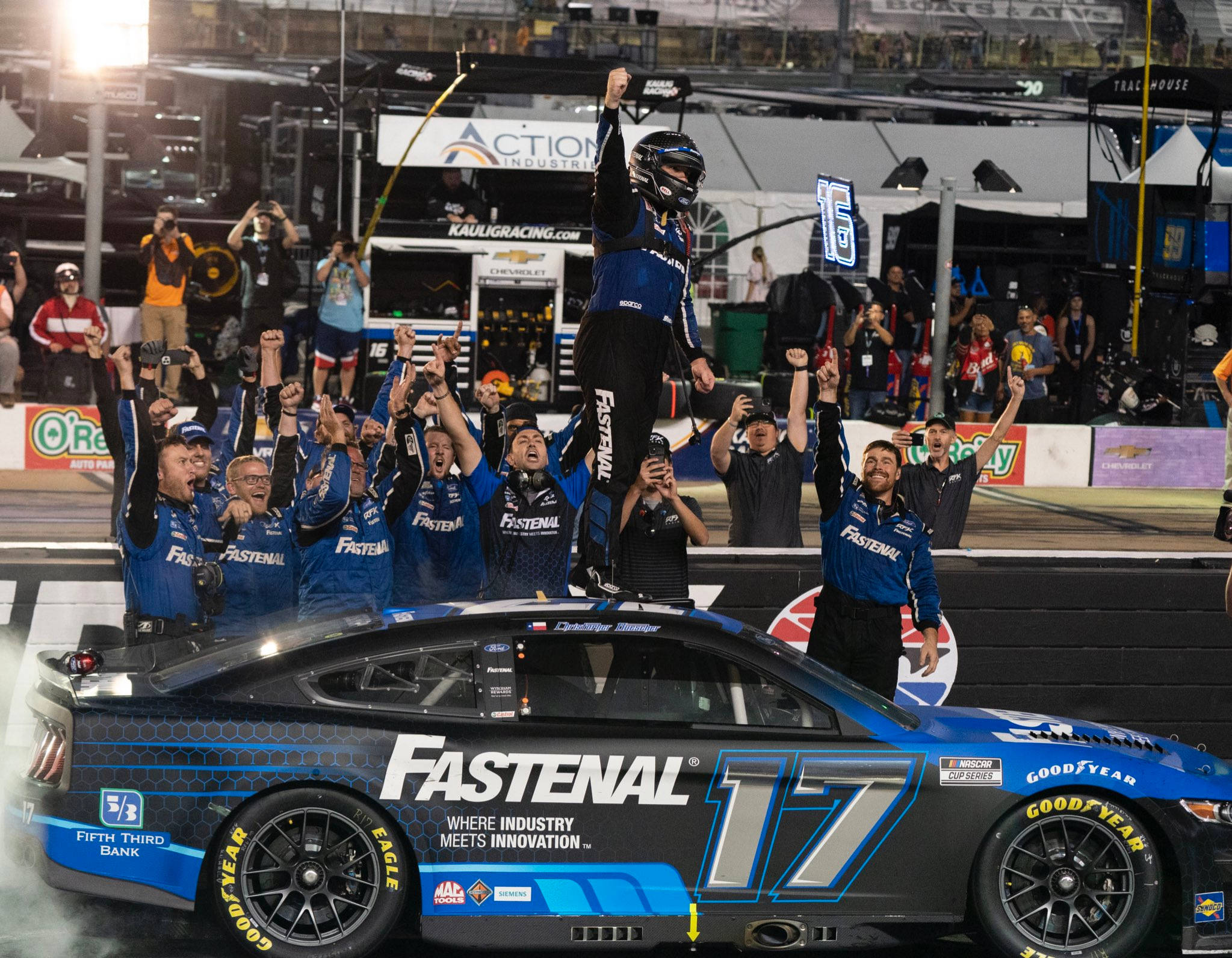 Racing 桌布 壁紙 — 一名身穿藍色裝備的NASCAR車手在夜間被他的團隊抬到他的藍黑#17 Fastenal賽車上，舉起拳頭慶祝勝利，背景是歡呼的粉絲。