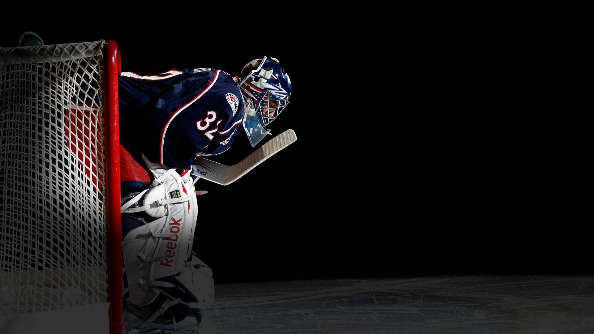 Columbus Blue Jackets 桌布 壁紙 — 一名身穿藍色裝備的冰球守門員在黑色背景的戲劇性聚光燈下蹲在他的球門旁。