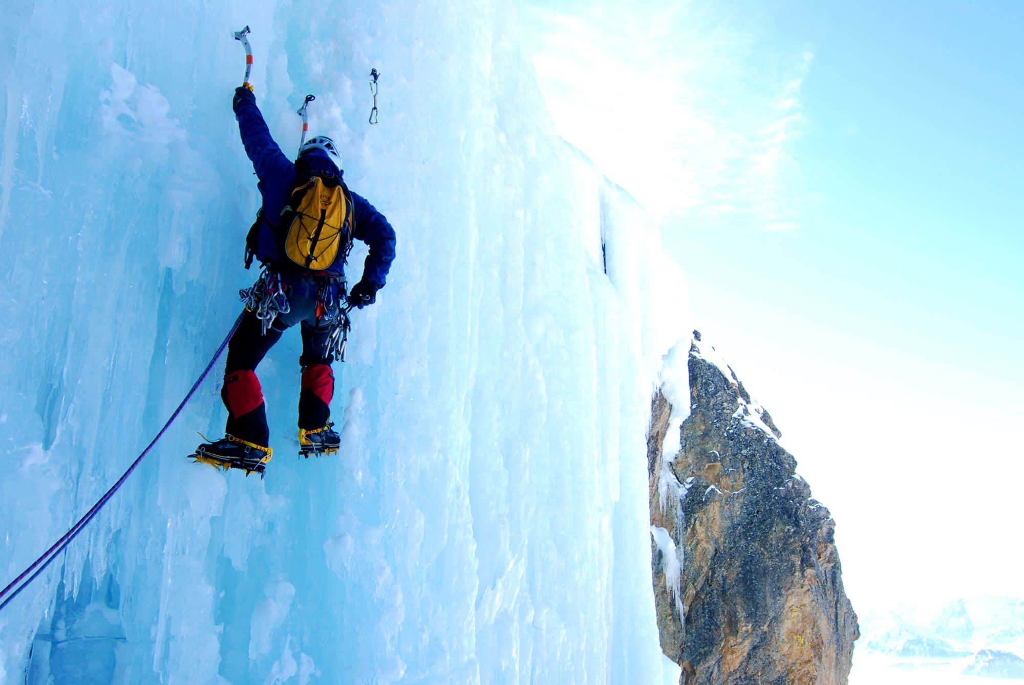 Ice Climbing 桌布 壁紙 — 一名身穿藍色裝備的攀岩者在明亮的藍天下攀登垂直的冰崖，附近有岩石露頭。