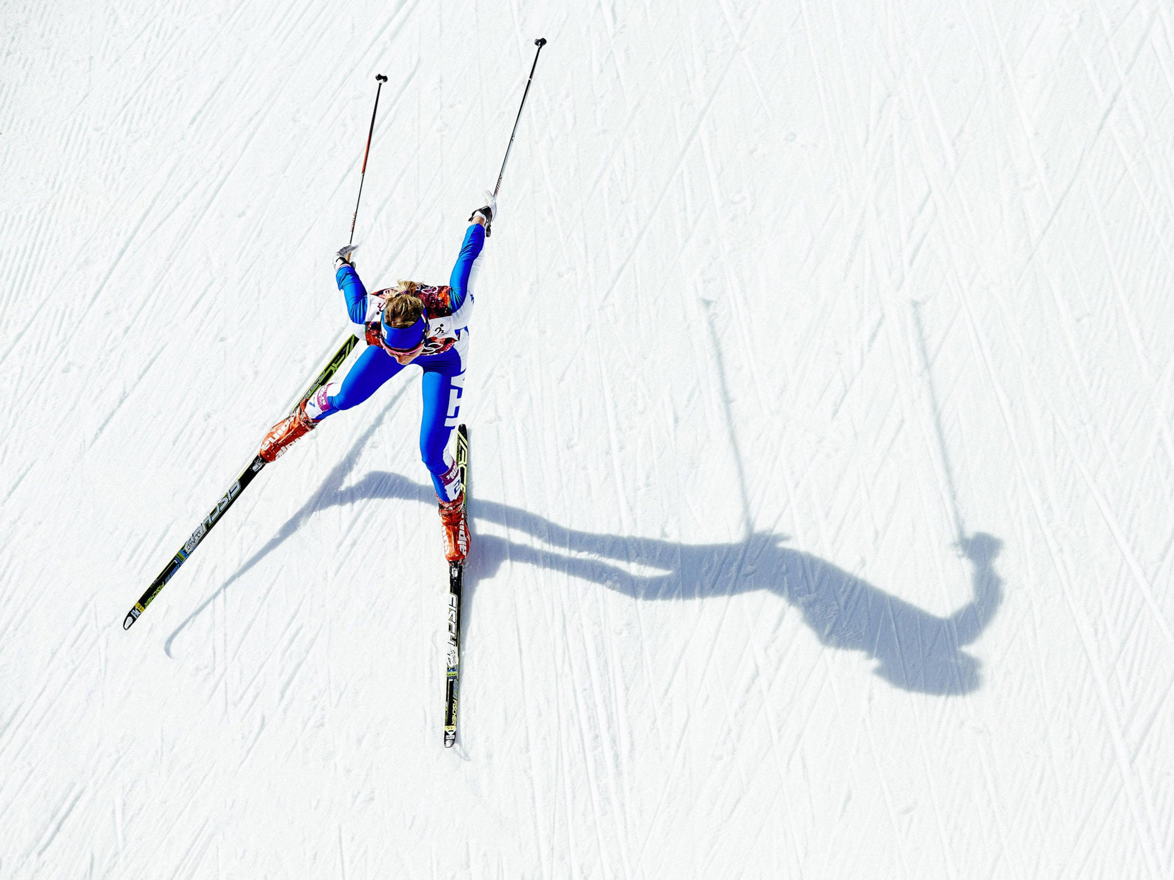 Winter Olympics 桌布 壁紙 — 一名身穿藍色裝備的滑雪者舉起雪杖，沿著雪坡滑下，在整齊的白色雪面上投下長長的影子。