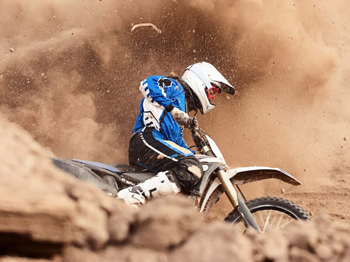 Dirtbike 桌布 壁紙 — 一名身穿藍色裝備的越野摩托車手在崎嶇的泥地上轉彎，揚起大片棕色塵土。