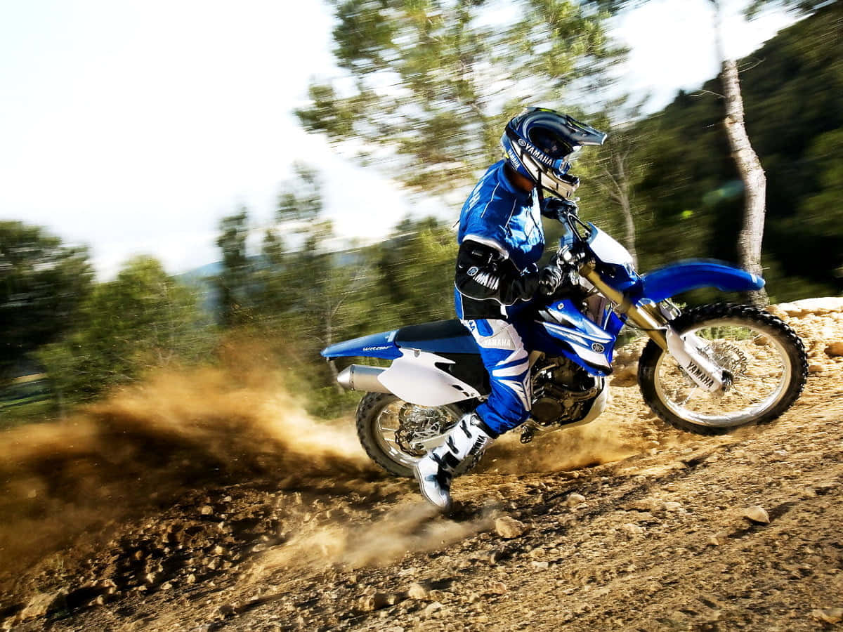 Motocross 桌布 壁紙 — 一名身穿藍色裝備的越野摩托車手在泥土小徑上傾斜過彎，揚起塵土，背景是模糊的森林。