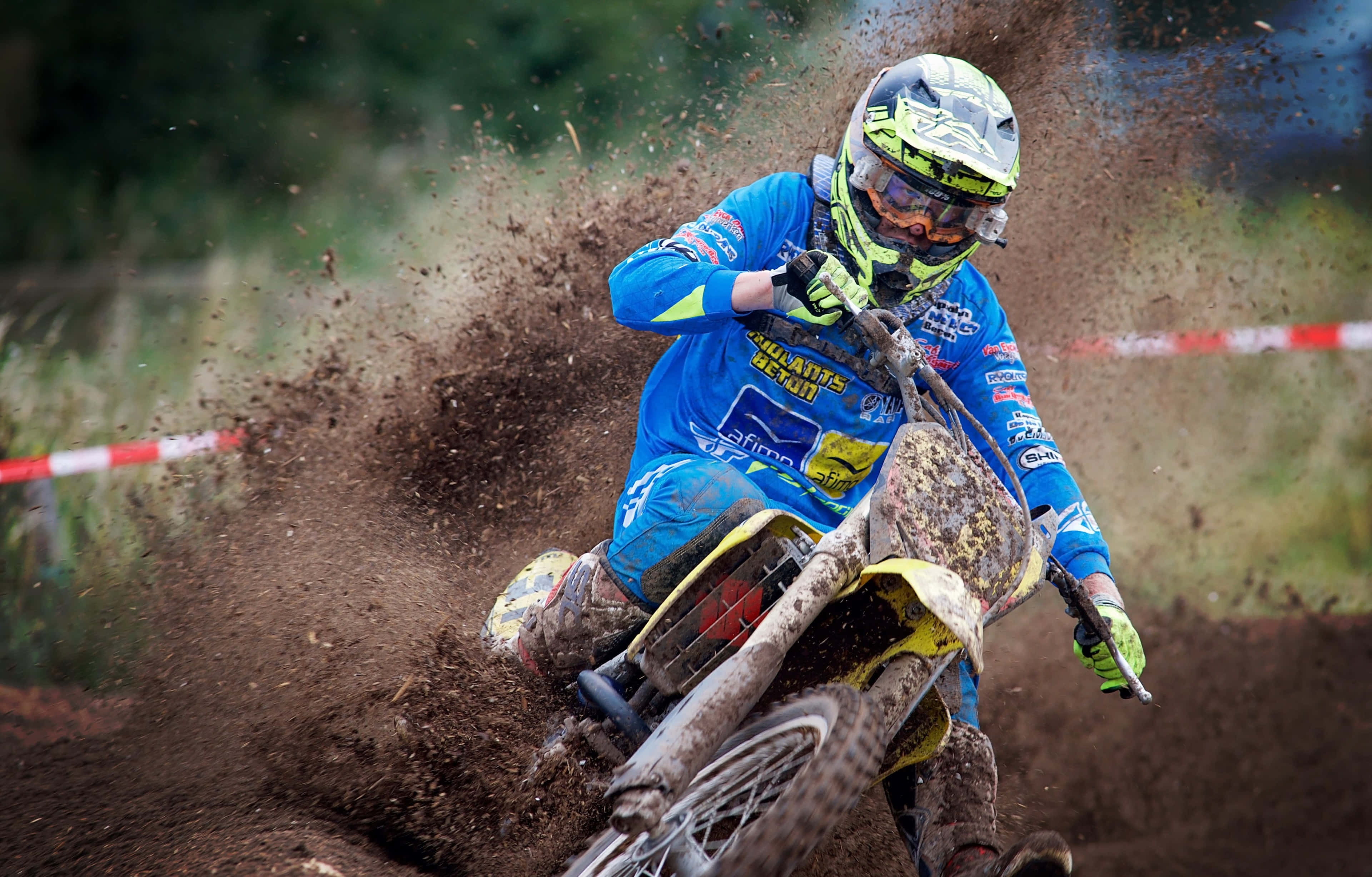 Motocross 桌布 壁紙 — 一名身穿藍色裝備的越野摩托車手在森林賽道上傾斜入泥濘彎道，揚起泥土。