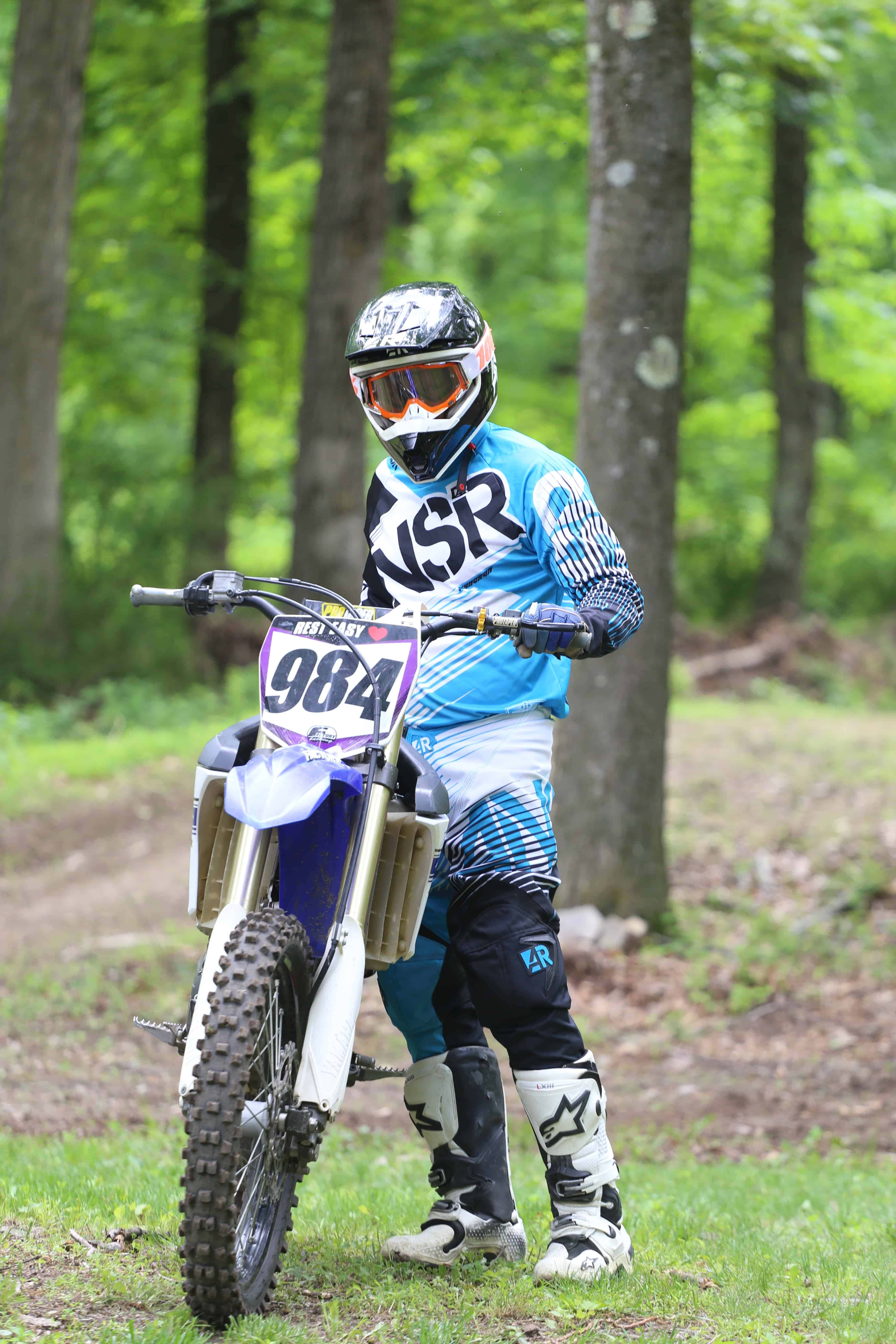 Dirtbike 桌布 壁紙 — 一名身穿藍色裝備的越野摩托車手站在陽光普照的森林空地中，旁邊是編號984的越野摩托車。