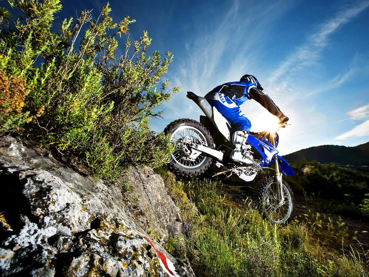 Motocross 桌布 壁紙 — 一名身穿藍色裝備的越野機車手，在陽光耀眼的明亮藍天下，於岩石小徑上傾身過急彎。