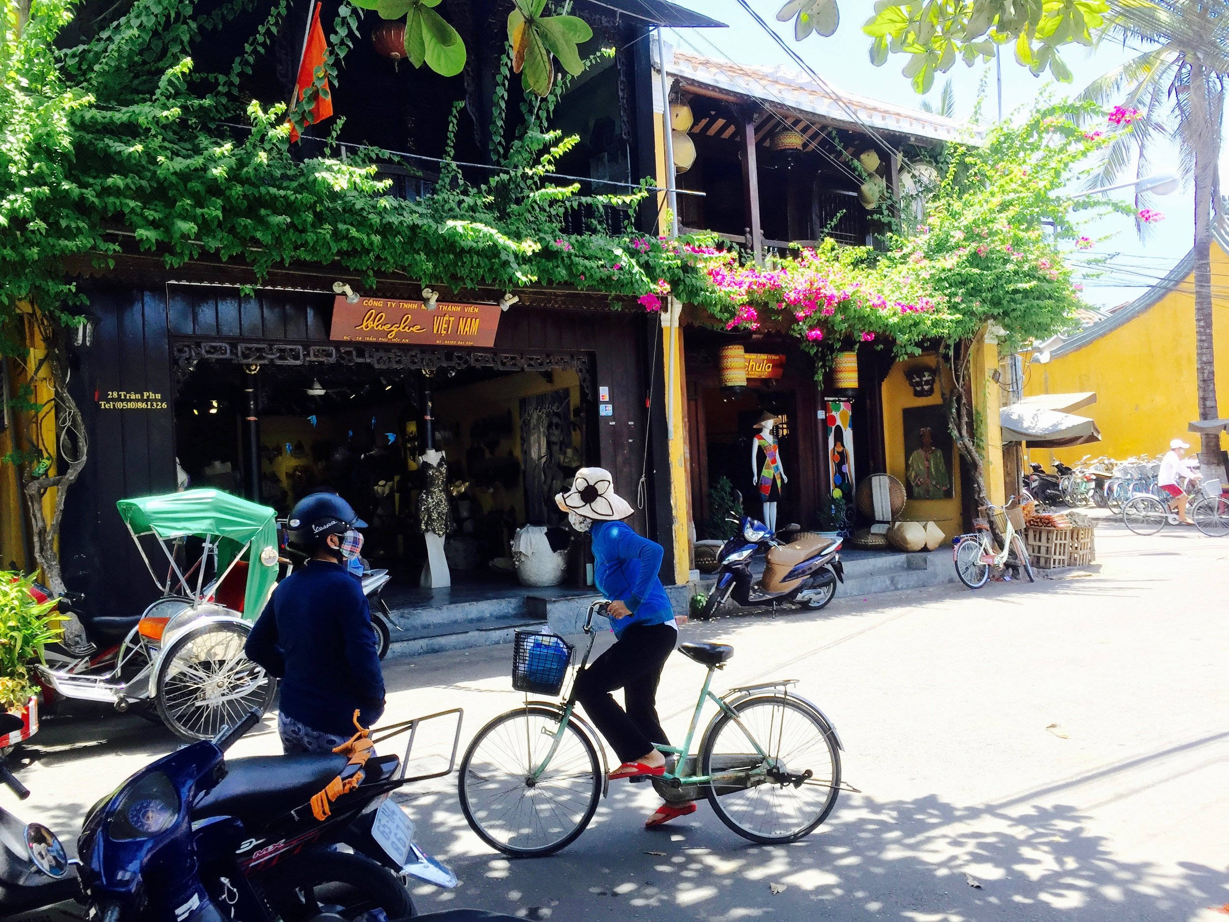 Hanoi 桌布 壁紙 — 一名身穿藍色襯衫、戴白色帽子的女子，踩著綠色腳踏車經過鮮黃色的建築物，建築物上點綴著茂盛的綠色植物和粉紅色的花朵，呈現陽光明媚的越南街景。
