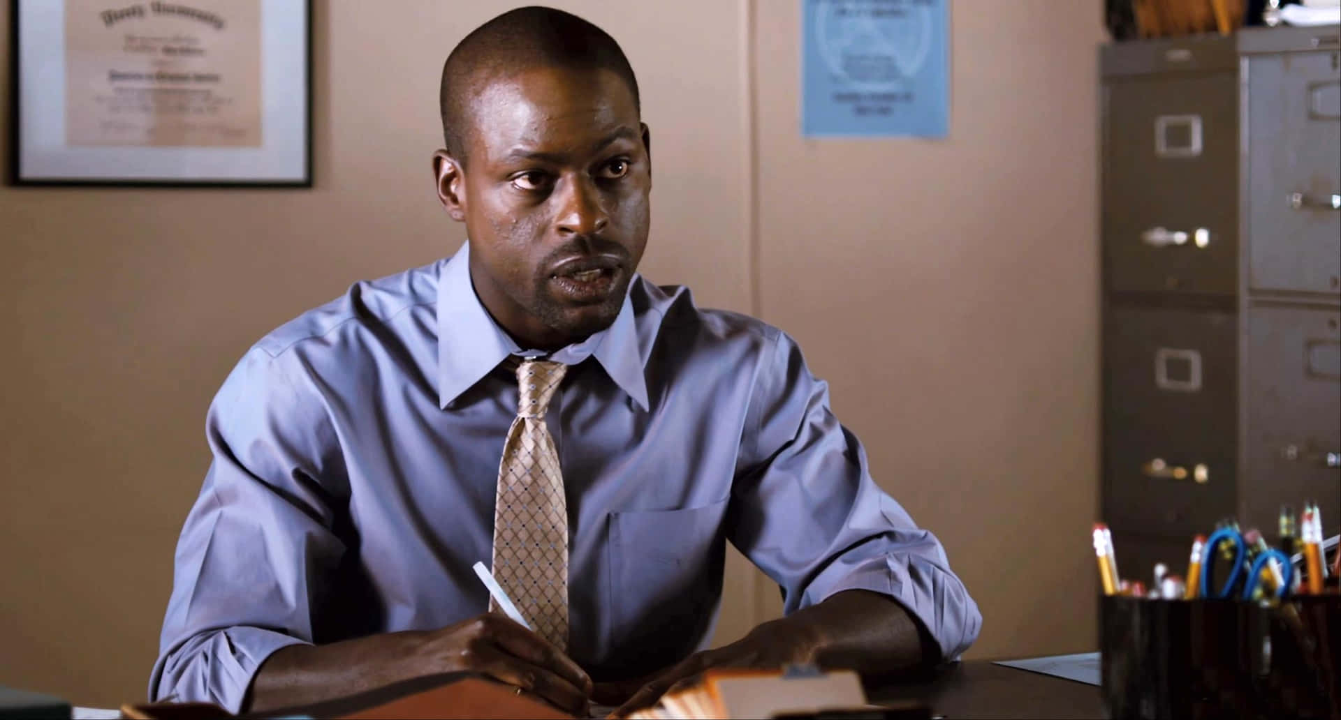 Sterling K Brown 桌布 壁紙 — 一名身穿藍色襯衫和圖案領帶的男子坐在辦公室的辦公桌前，手持鋼筆，表情嚴肅。