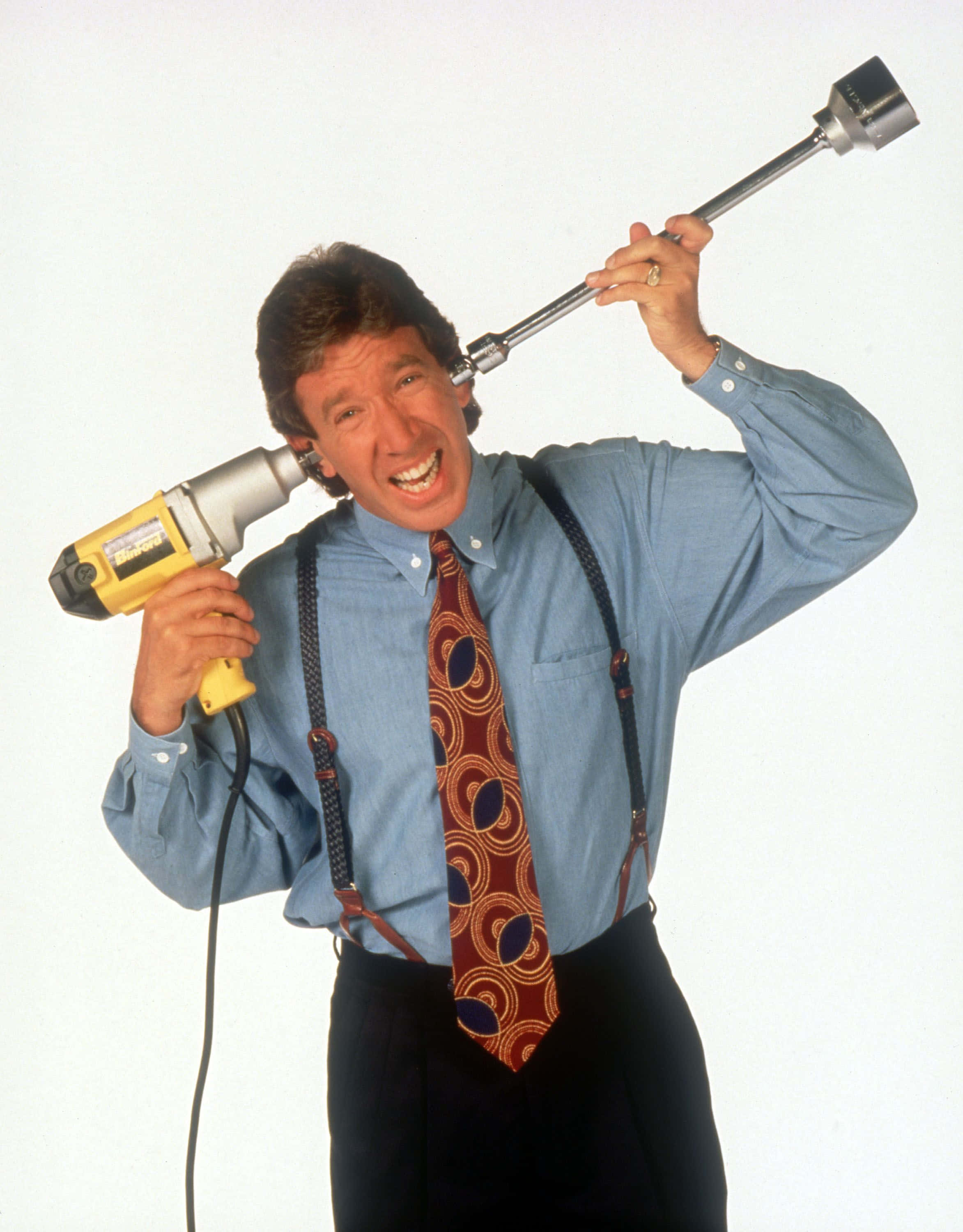Tim Allen 桌布 壁紙 — 一名身穿藍色襯衫和圖案領帶的男子，在純白色背景前，將黃色電鑽像麥克風一樣舉起，咧嘴而笑。