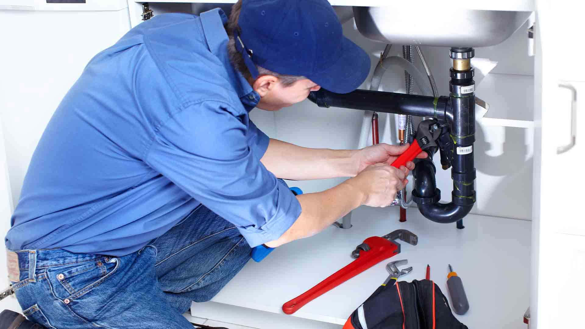 Plumbing 桌布 壁紙 — 一名身穿藍色襯衫和帽子的水管工用紅色鉗子修理白色廚房水槽下方的管道。