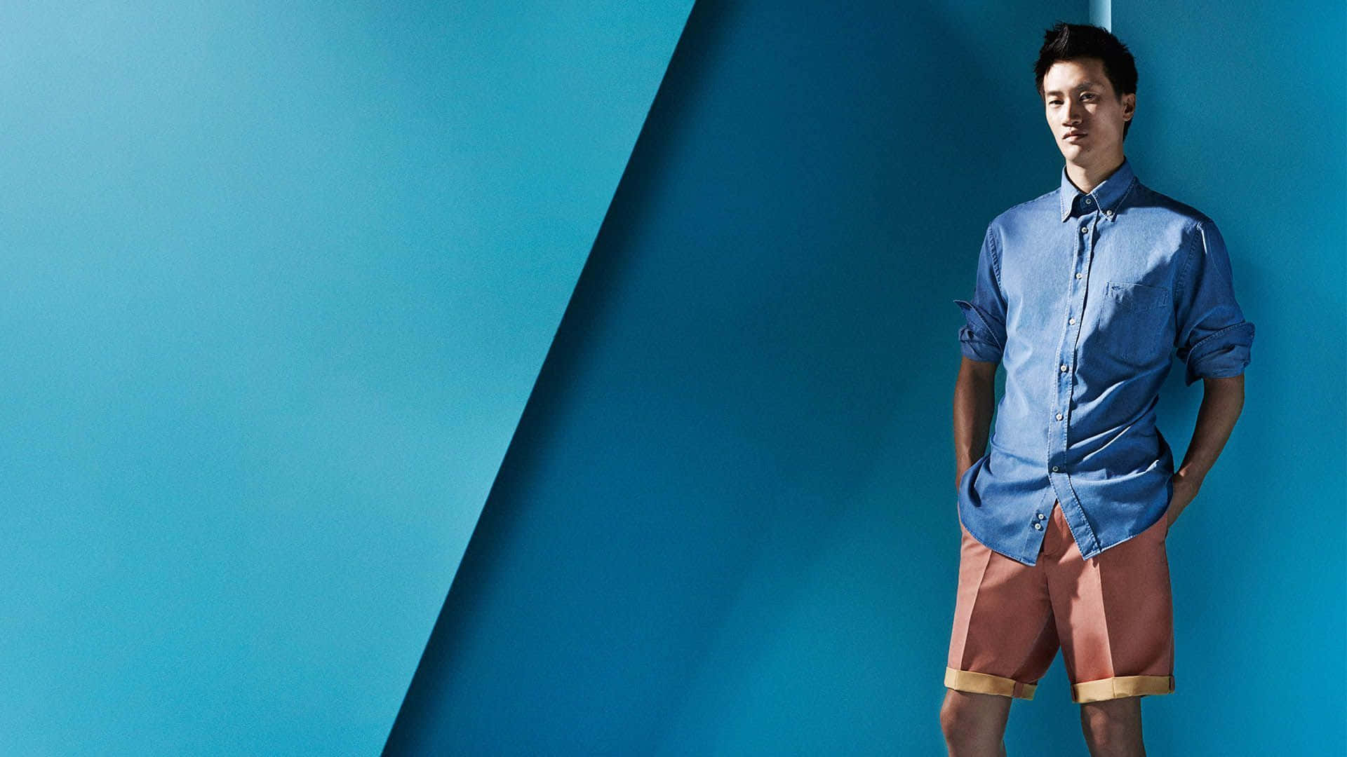 Mens Fashion 桌布 壁紙 — 一名身穿藍色襯衫和珊瑚色短褲的男子自信地站在大膽的藍色幾何牆前。