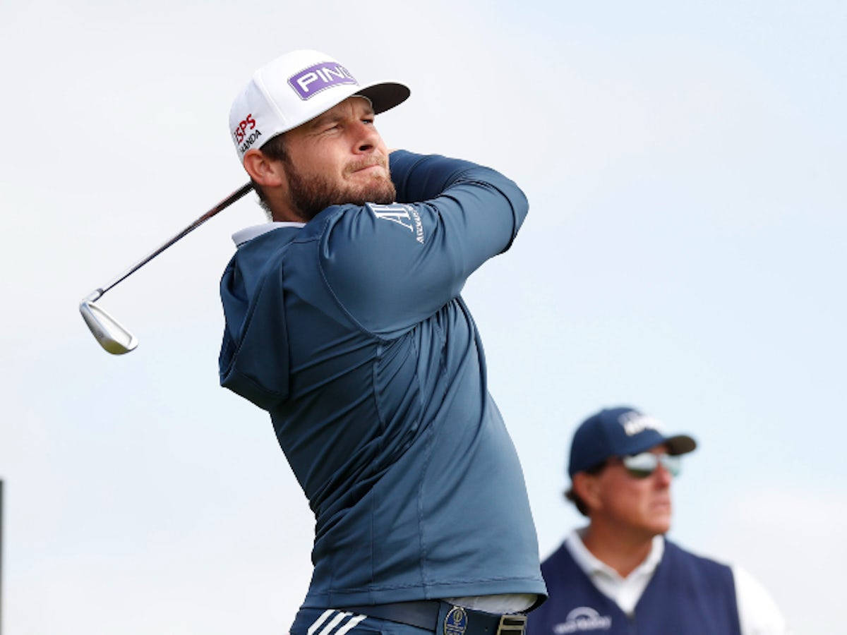 Tyrrell Hatton 桌布 壁紙 — 一名身穿藍色襯衫和白色帽子的男性高爾夫球手，在明亮的天空下專注地揮桿。