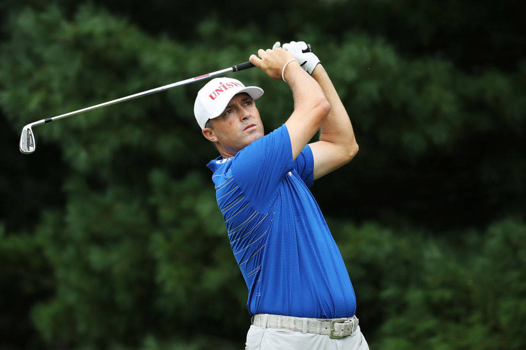 Ryan Palmer 桌布 壁紙 — 一名身穿藍色襯衫和白色帽子的男性高爾夫球手，在模糊的綠色森林背景下執行強力揮桿。