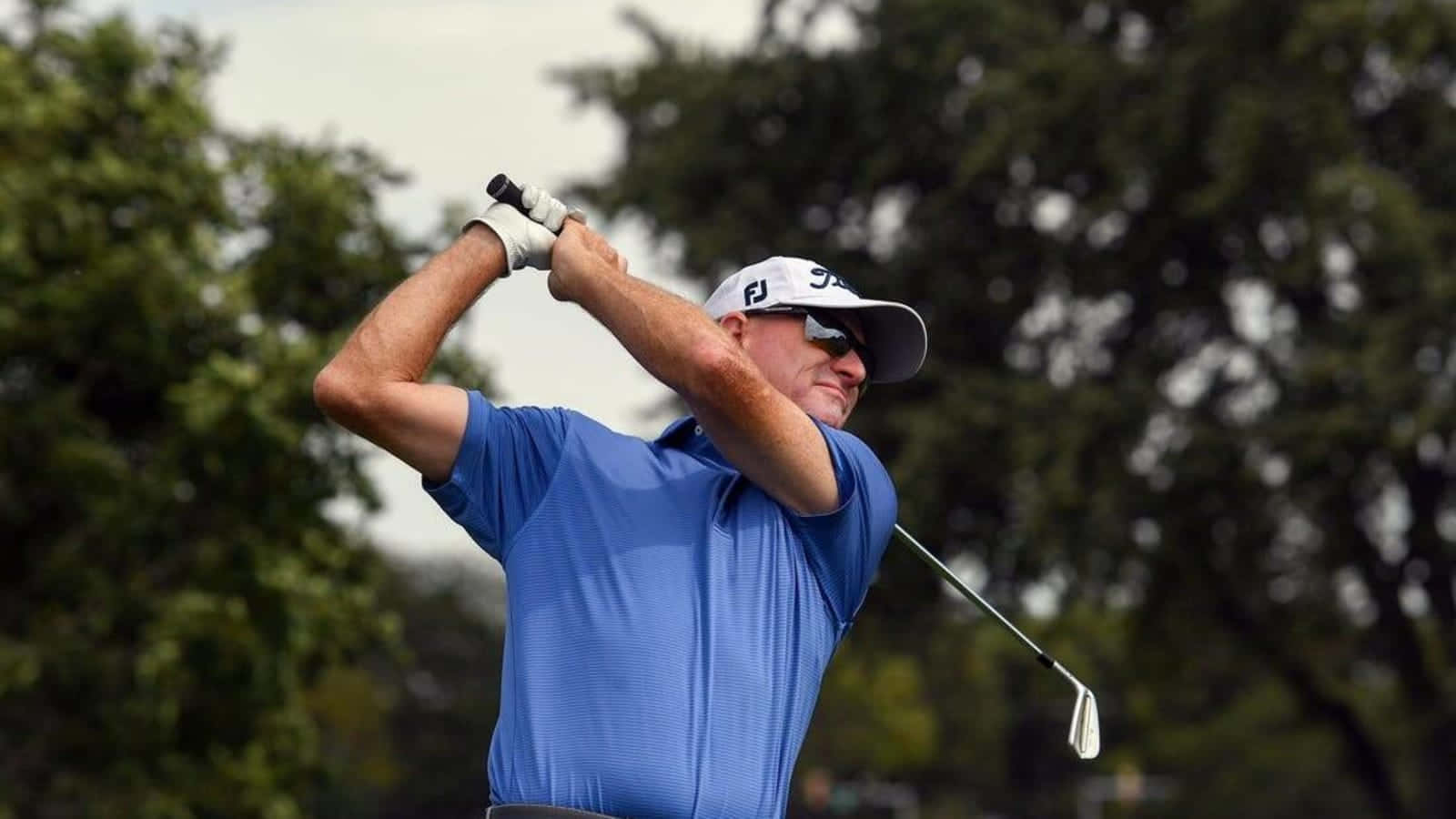  壁紙 — 一名身穿藍色襯衫和白色帽子的高爾夫球手，在茂密綠樹的背景下，專注地揮桿。
