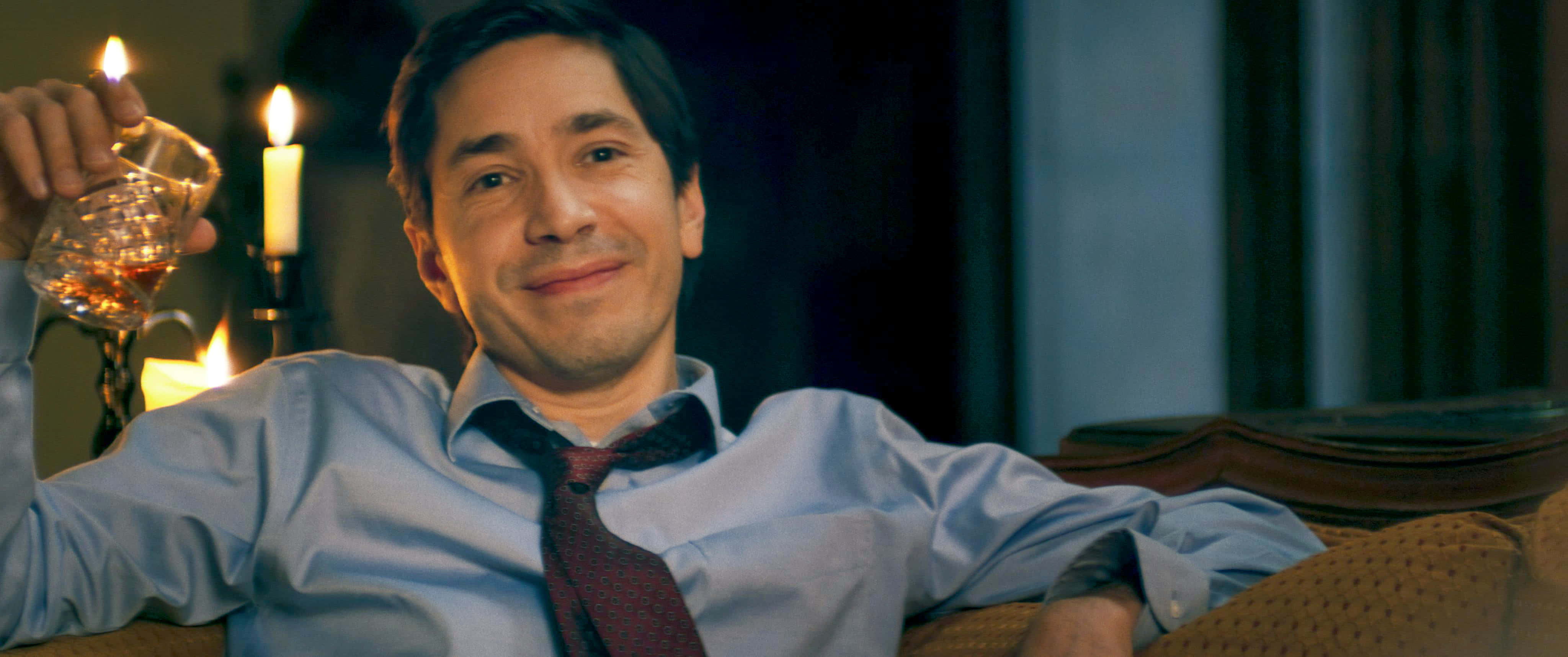 Justin Long 桌布 壁紙 — 一名身穿藍色襯衫和領帶的男子，在燭光昏暗的房間裡拿著一杯威士忌微笑。