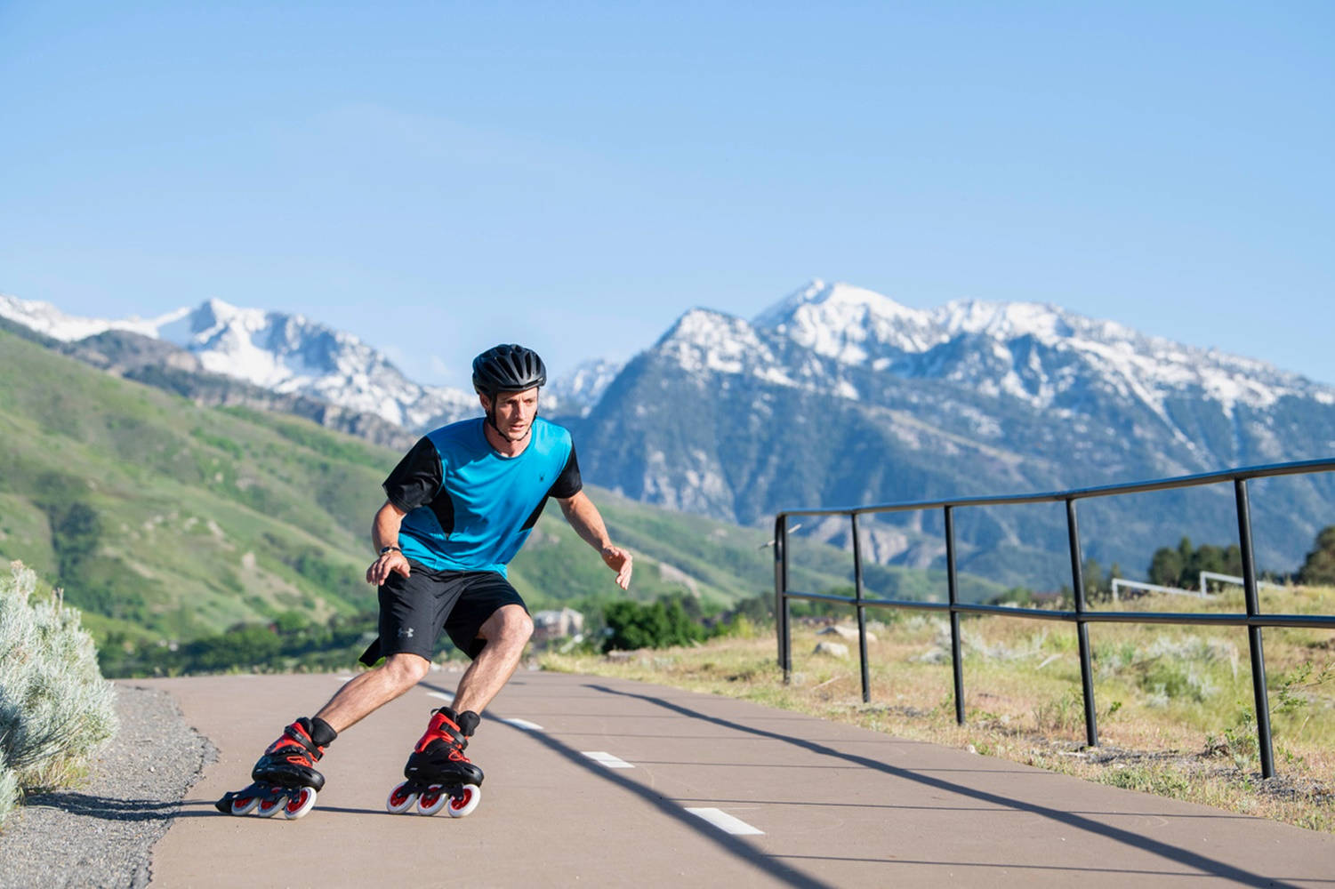 skating 桌布 壁紙 — 一名身穿藍色襯衫和頭盔的男子在鋪砌的小徑上溜冰，背景是白雪覆蓋的山脈和綠色山丘，天空清澈湛藍。