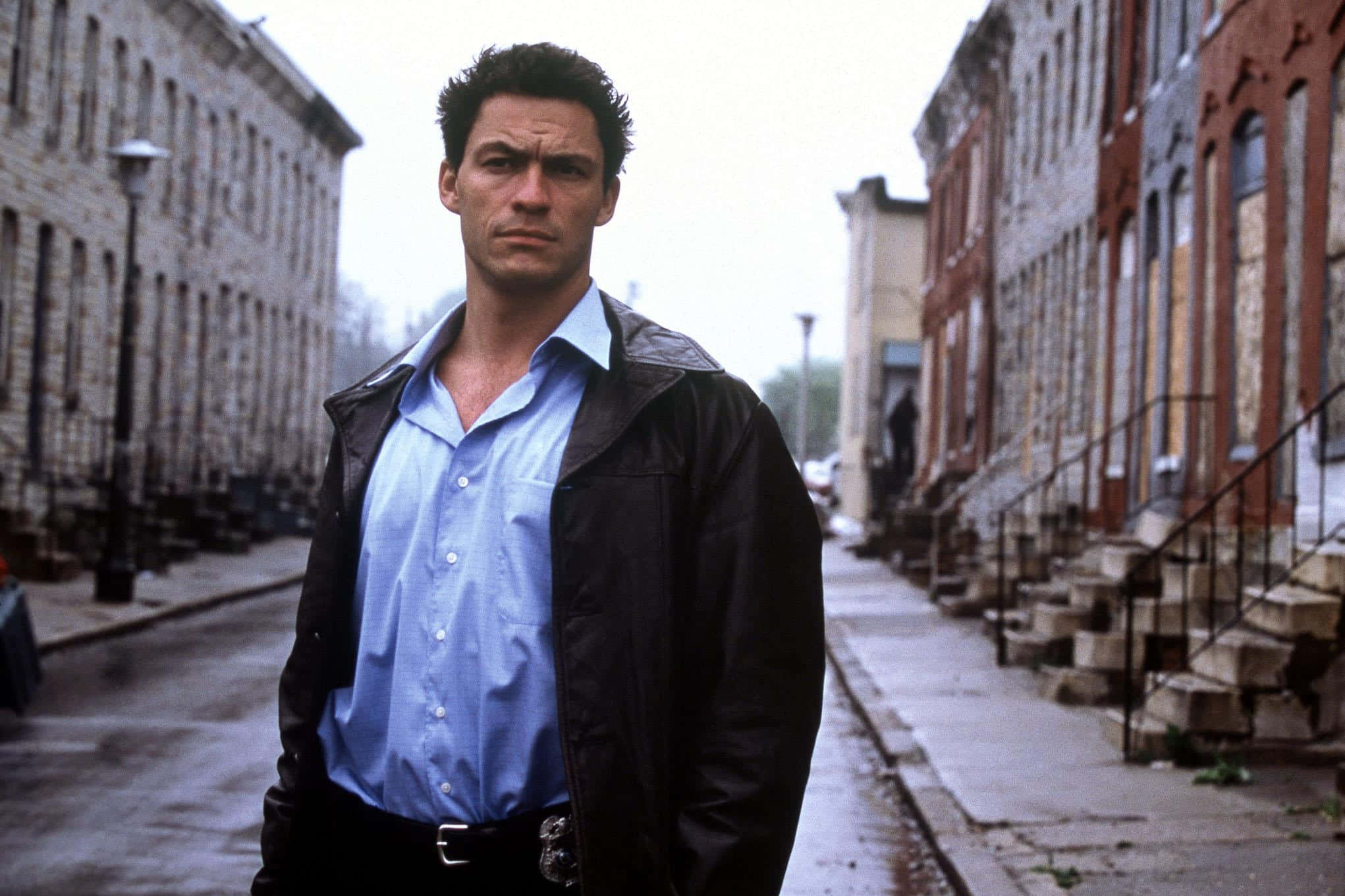 Dominic West 桌布 壁紙 — 一名身穿藍色襯衫和黑色夾克的男子，在陰天之下，莊嚴地站在一條濕漉漉的都市街道上，兩旁是磚造連棟房屋。