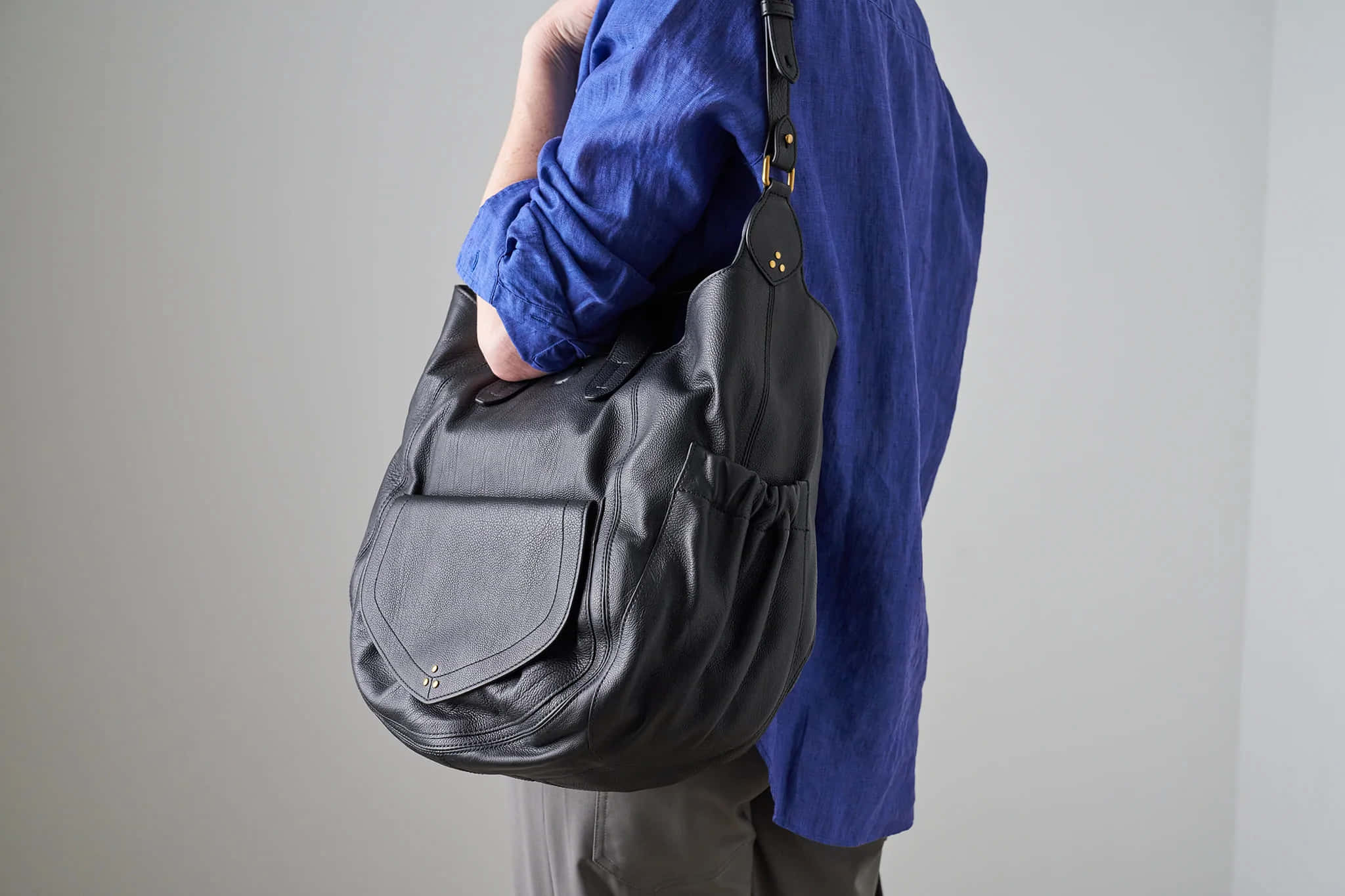 Handbag 桌布 壁紙 — 一名身穿藍色襯衫的人，在純灰色牆壁前，拿著一個帶有金色金屬配件的黑色皮革流浪包。