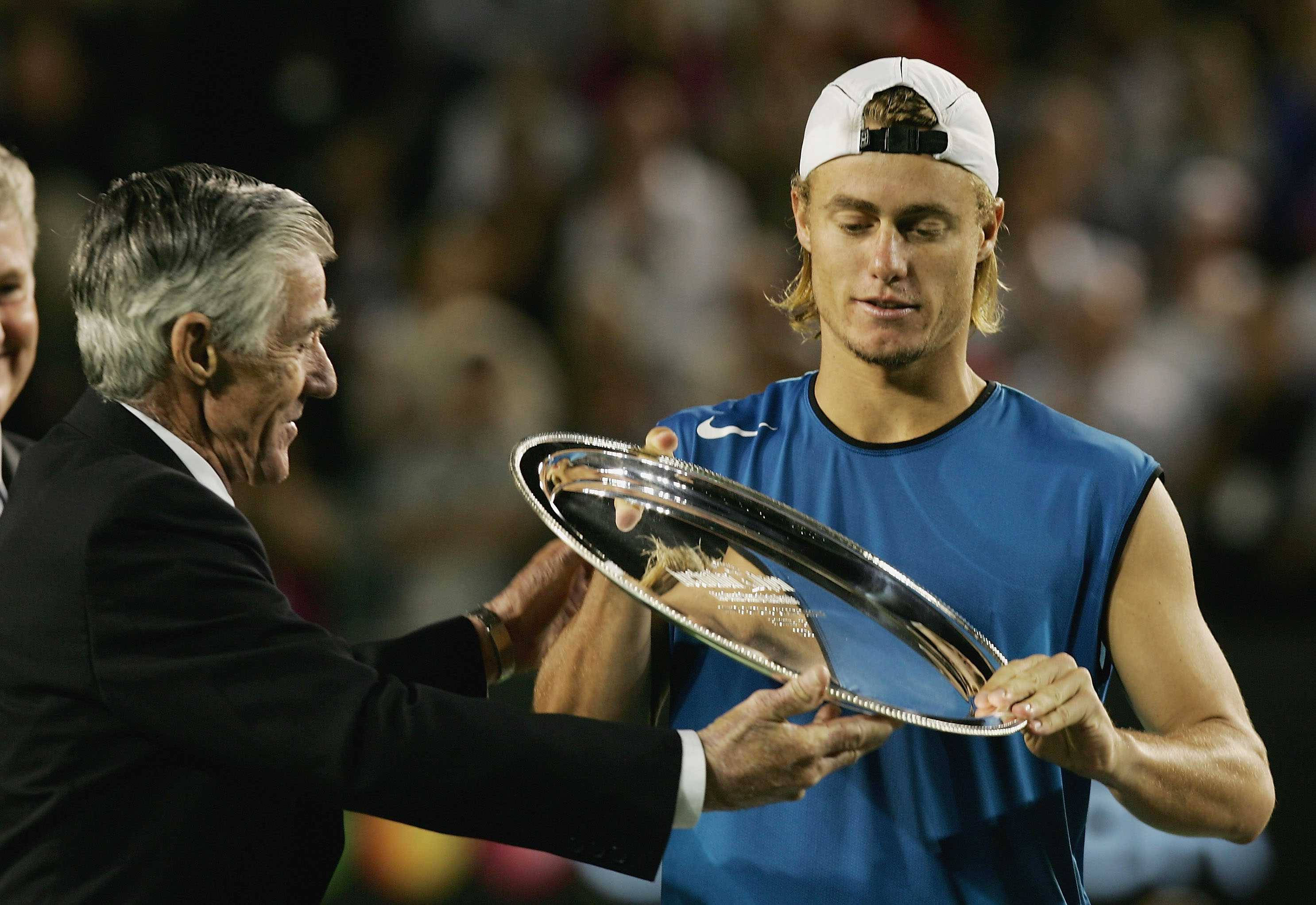 Australian Open 桌布 壁紙 — 一名身穿藍色襯衫的年輕網球選手，在體育場燈光下，從一位身穿西裝的年長男子手中接過一個大型銀色獎盃。