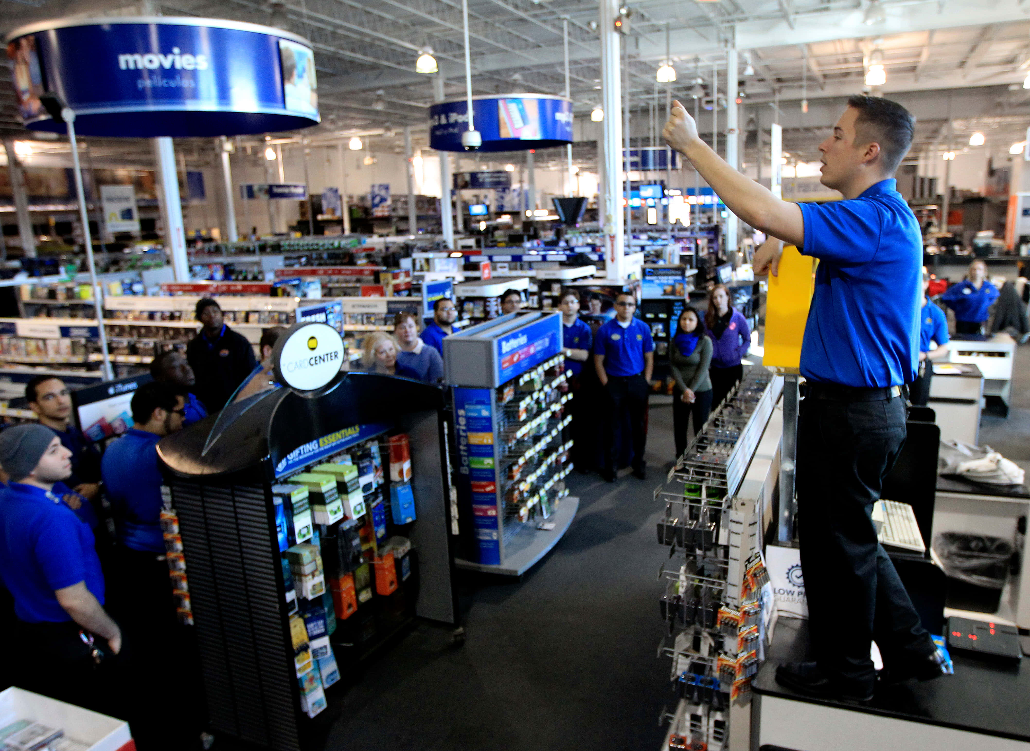 Best Buy 桌布 壁紙 — 一名身穿藍色襯衫的店員站在收銀台上，向一群同事做手勢，店內光線明亮。