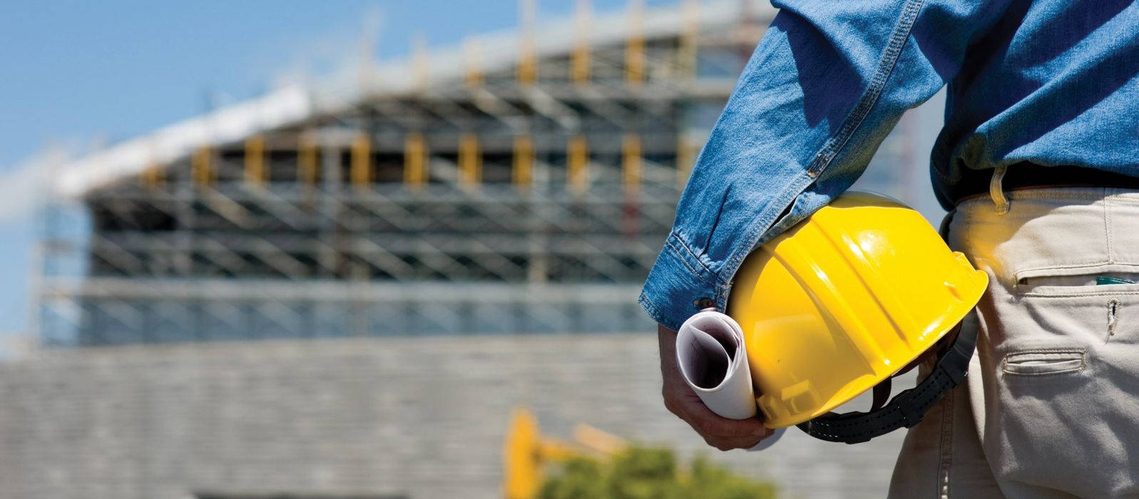 Work 桌布 壁紙 — 一名身穿藍色襯衫的建築工人，手持黃色安全帽和捲起的藍圖，背景是一座搭有鷹架的大型建築物。
