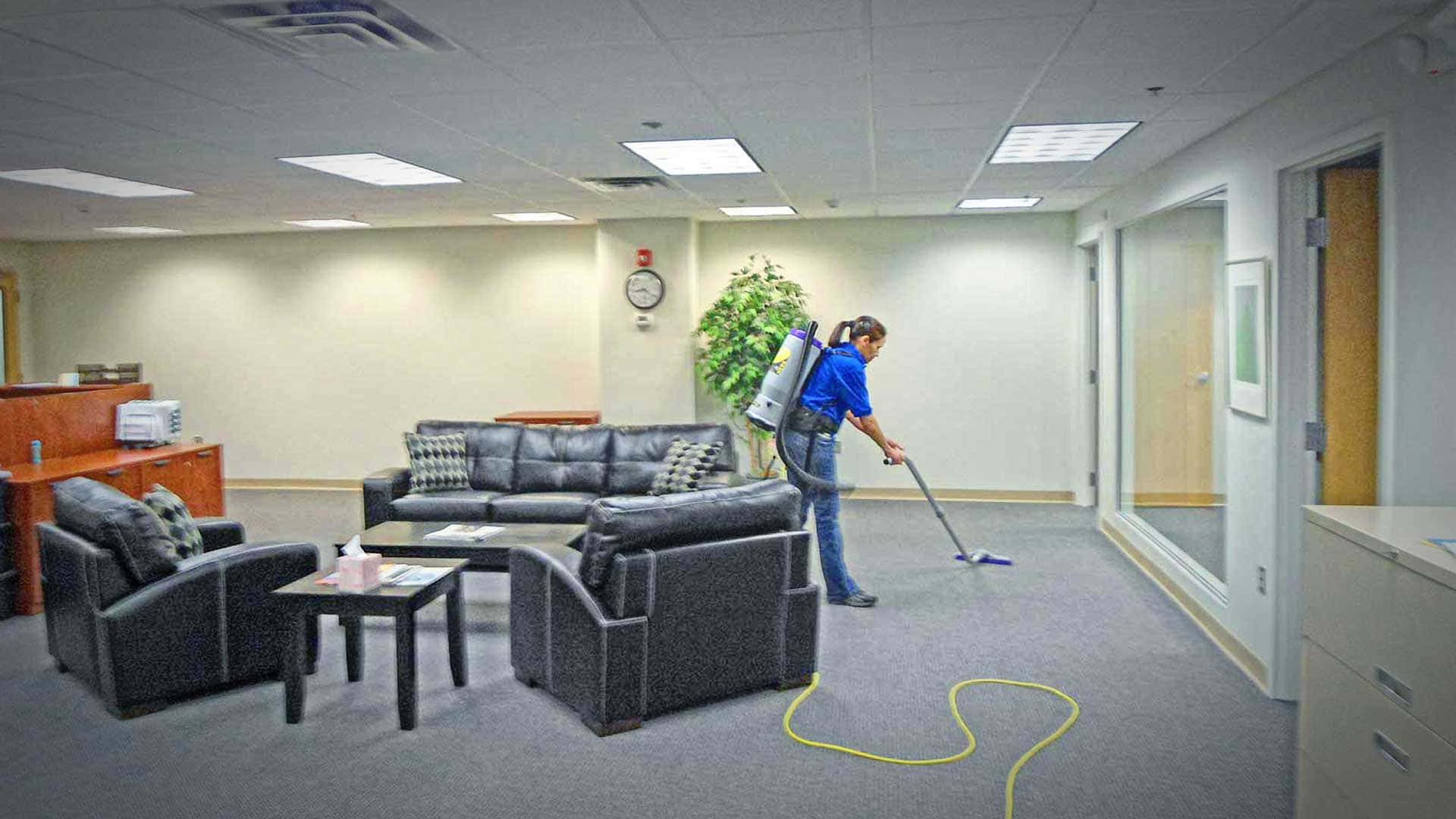 Cleaning 桌布 壁紙 — 一名身穿藍色襯衫的清潔工正在用吸塵器清潔鋪有地毯的辦公室大廳，大廳內有黑色皮革沙發和盆栽植物。