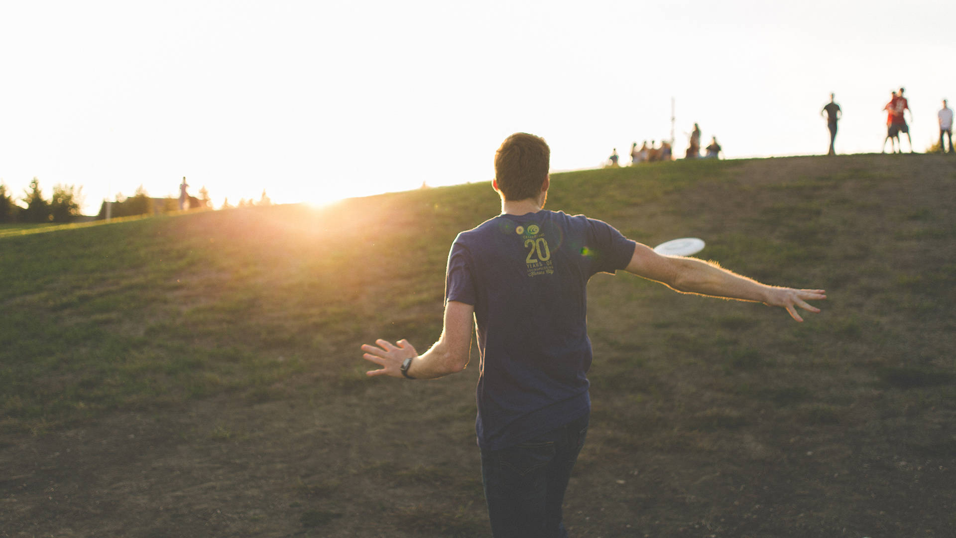 Ultimate Frisbee 桌布 壁紙 — 一名身穿藍色襯衫的男子在日落時分將飛盤扔過草地山坡，遠處有其他人觀看。