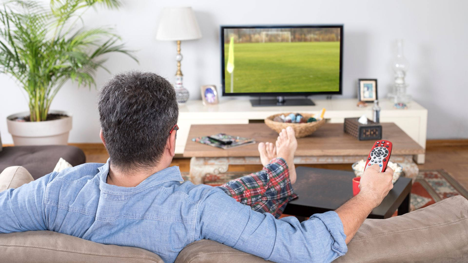 tv 桌布 壁紙 — 一名身穿藍色襯衫的男子懶洋洋地躺在沙發上，手持紅色遙控器，觀看電視上的足球比賽。