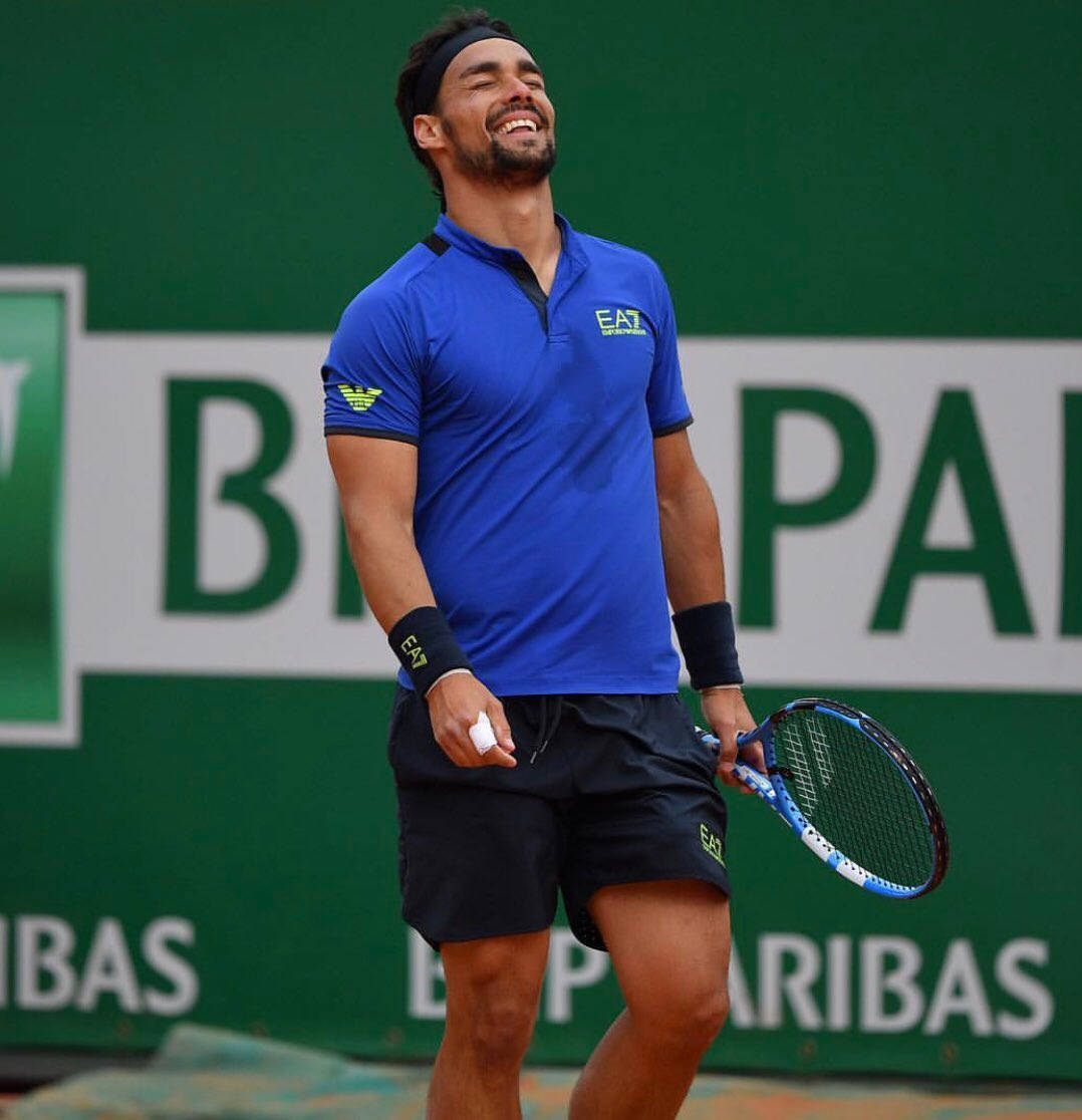 Tennis 桌布 壁紙 — 一名身穿藍色襯衫的男性網球選手在紅土球場上開心地笑著，手持球拍，身後有綠色廣告牆。