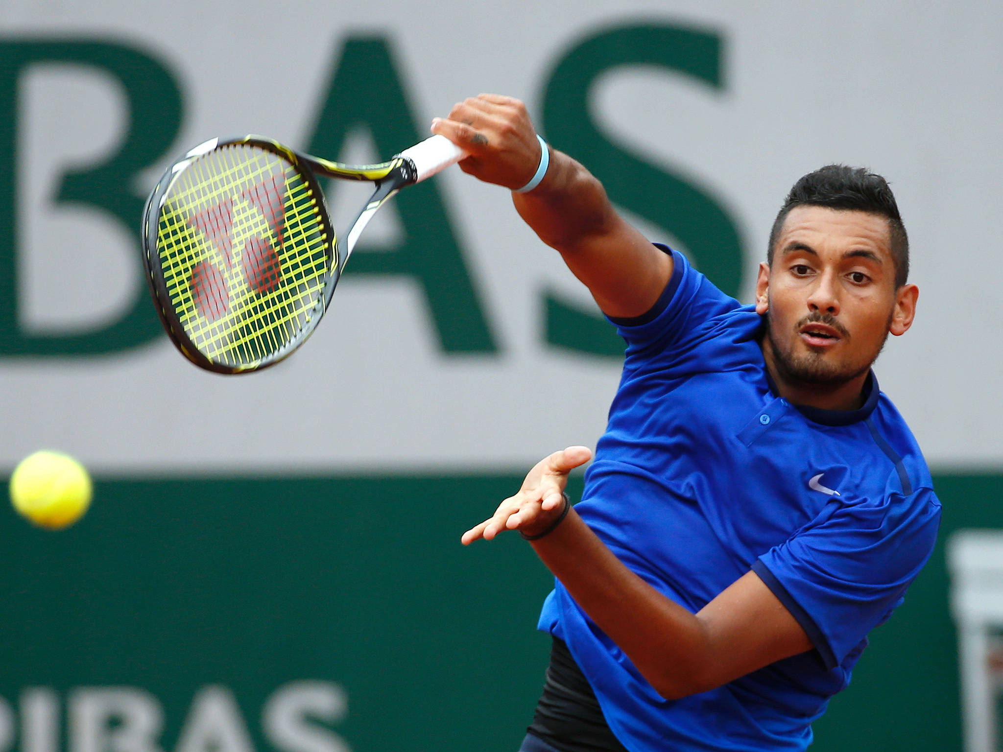 Kyrgios 桌布 壁紙 — 一名身穿藍色襯衫的男網球選手在紅土球場比賽中揮拍擊打黃色網球。