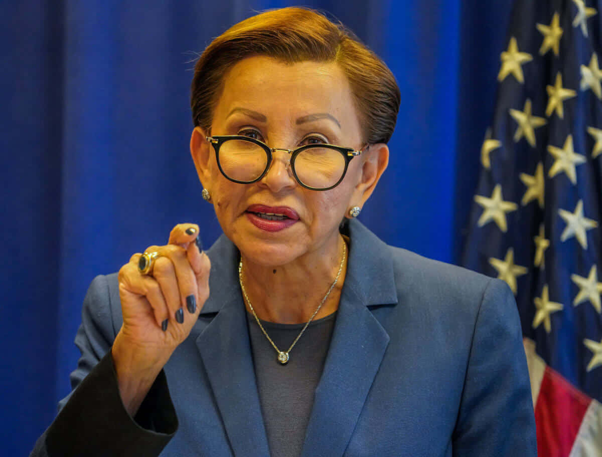 pointing 桌布 壁紙 — 一名身穿藍色西裝、戴眼鏡的女性在藍色窗簾和美國國旗前說話時做手勢。